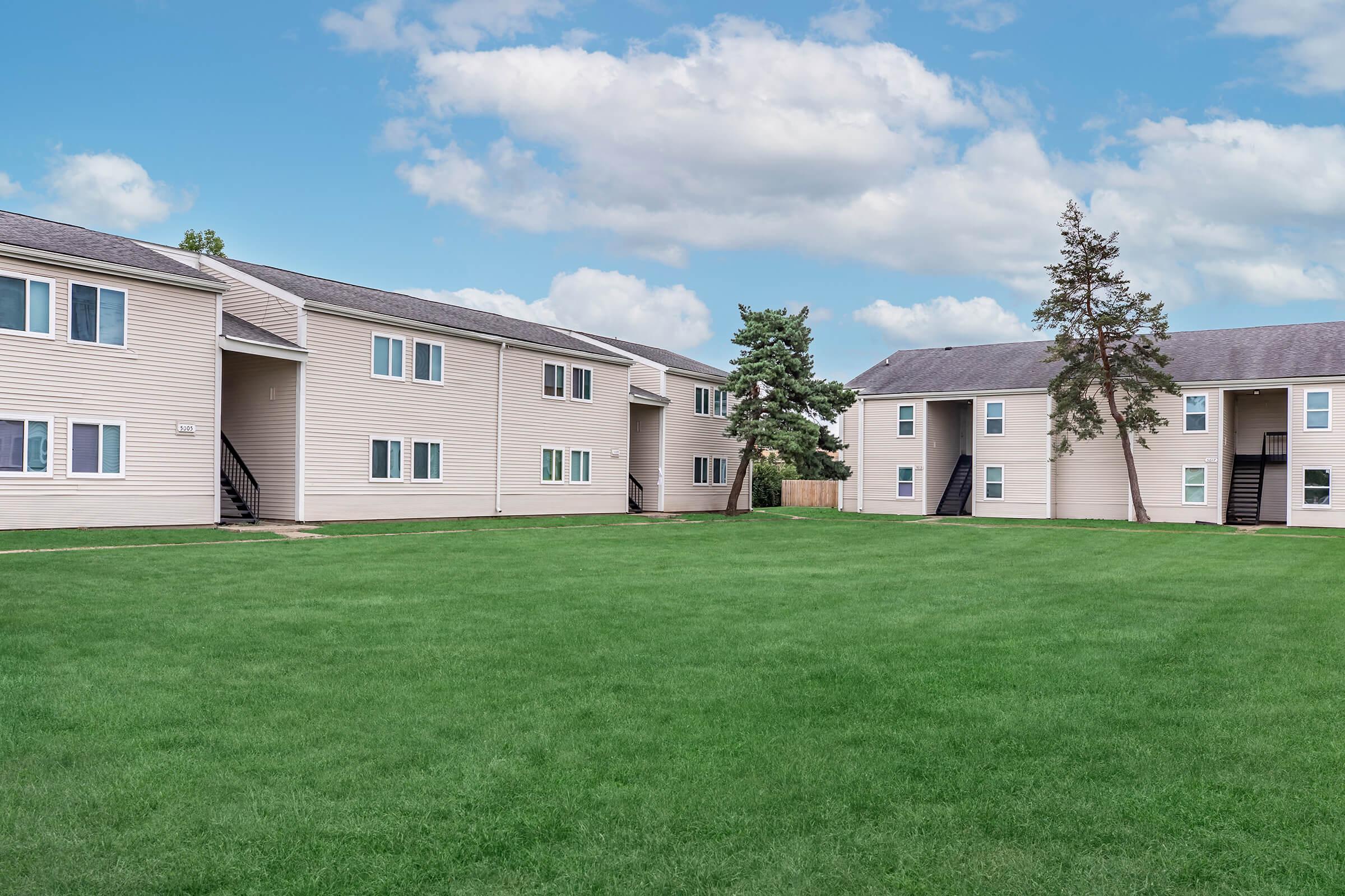 A neatly maintained residential area featuring several two-story apartment buildings made of light-colored siding. The buildings surround a wide, lush green lawn, with clear blue skies and fluffy white clouds in the background. Trees are visible near the buildings, contributing to the pleasant outdoor atmosphere.