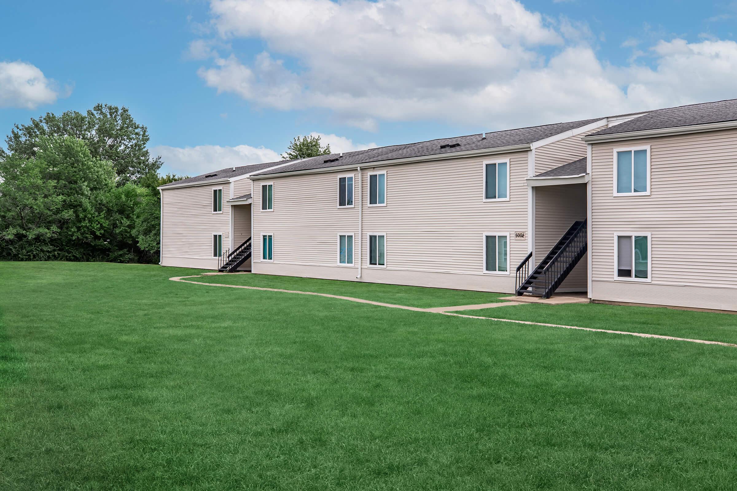 A view of a multi-unit residential building with a light-colored exterior, surrounded by a green lawn. The building features multiple windows and staircases leading to individual units. A pathway runs through the grass, providing access to the entrance of the building.