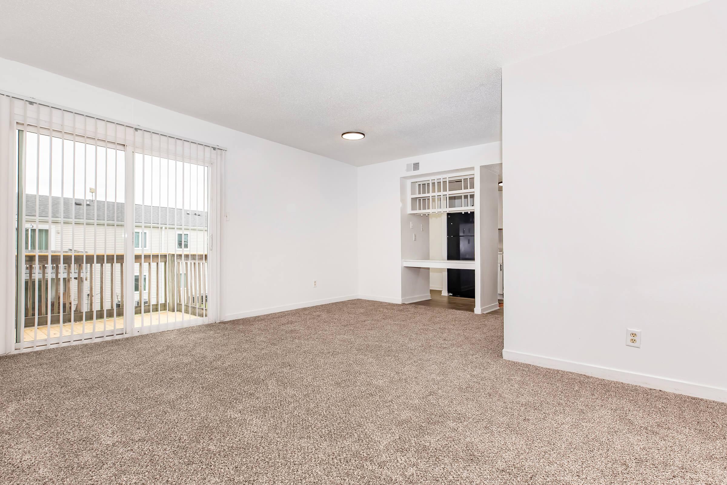Spacious, empty living room with light-colored carpet, large windows with vertical blinds letting in natural light, and an open view into a kitchen area with black appliances. The walls are painted a light color, and there is a ceiling light fixture.