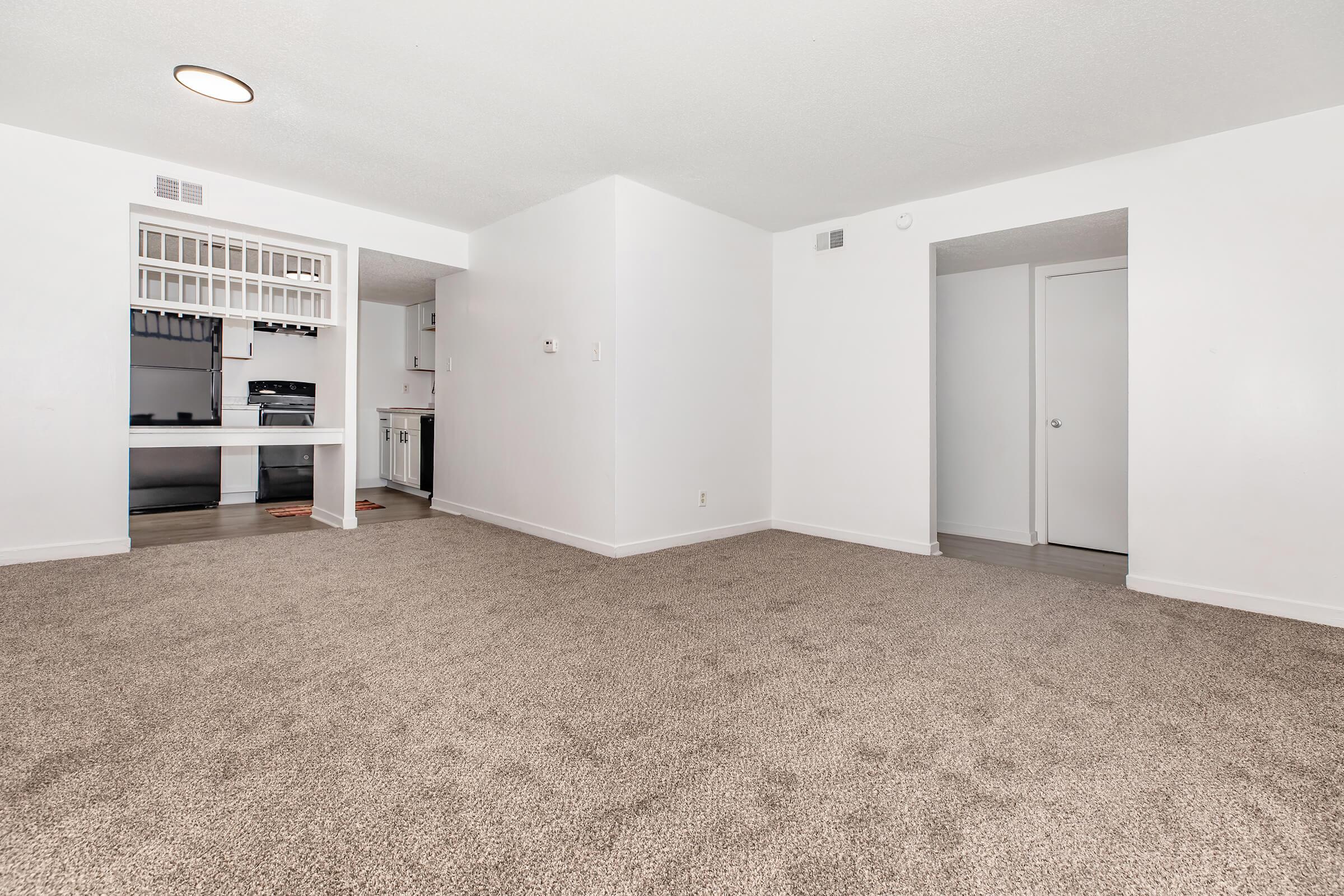 A spacious, empty living area with light-colored walls and tan carpeting. The room features an open layout with a small kitchen visible in the background, equipped with dark appliances and cabinetry. Two doorways on the right lead to other rooms, and there is a ceiling light fixture above.