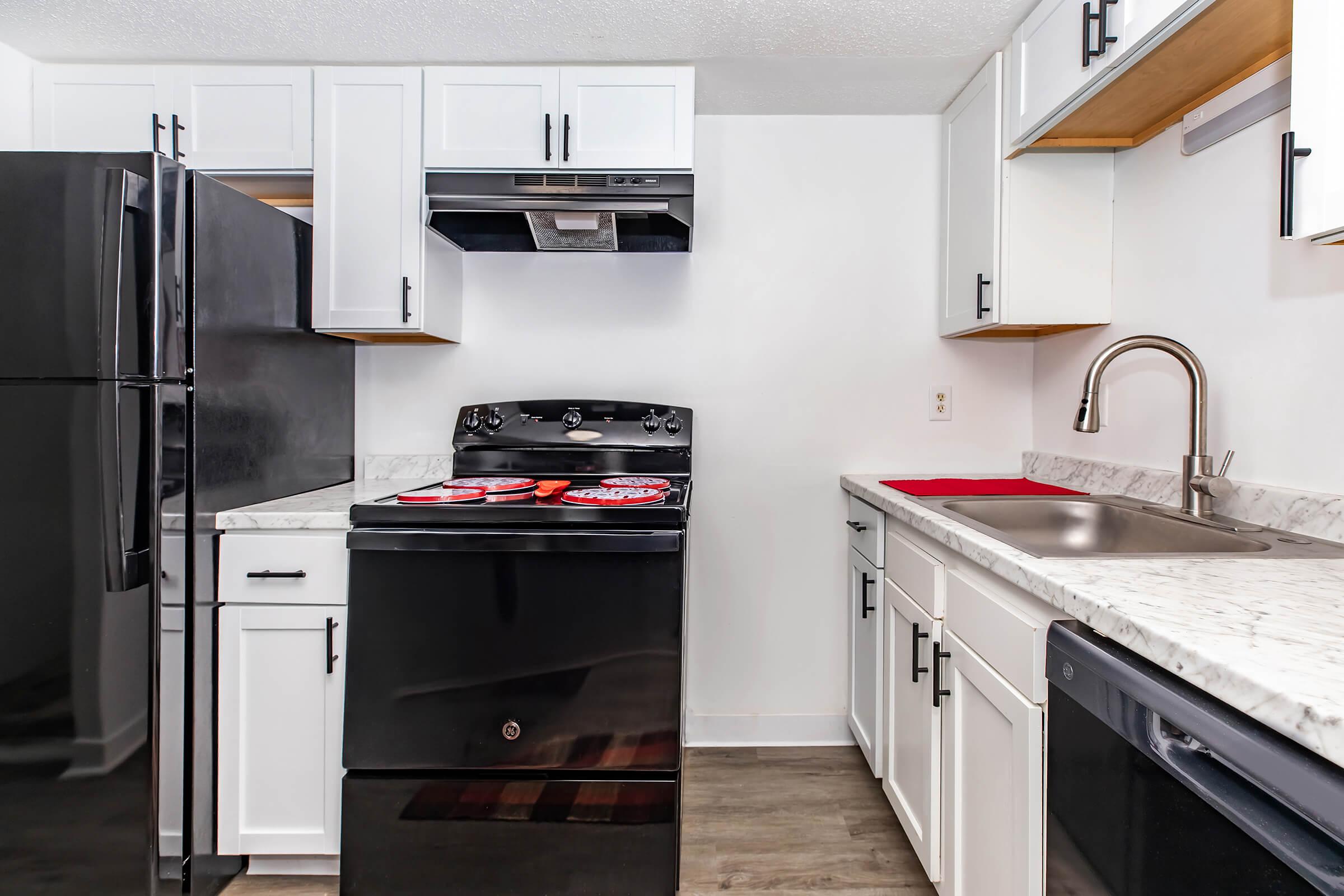 Modern kitchen featuring a black refrigerator and black stove with a red tray on top. White cabinets with wooden shelves and a stainless steel sink are visible, along with a black dishwasher. The countertop is made of light-colored marble, and the flooring is a neutral wood tone.