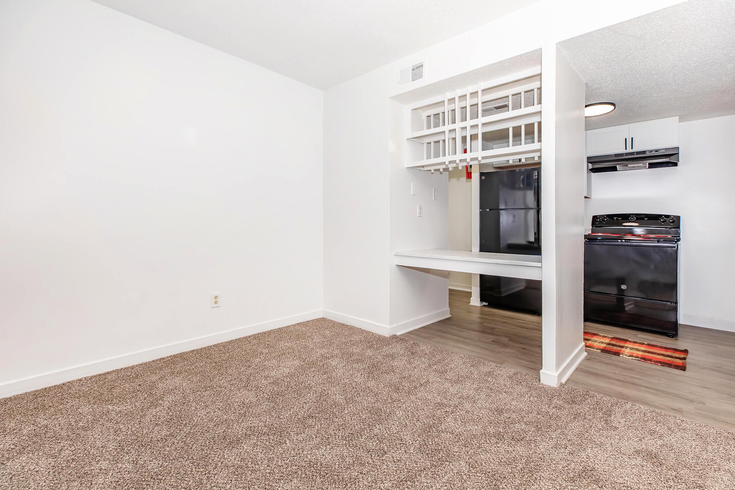 A clean and modern living space with light-colored walls and a beige carpet. An open kitchen area is visible, featuring black appliances, including a refrigerator, stove, and overhead cabinets. A small dining counter separates the kitchen from the living area, creating a cozy and functional layout.