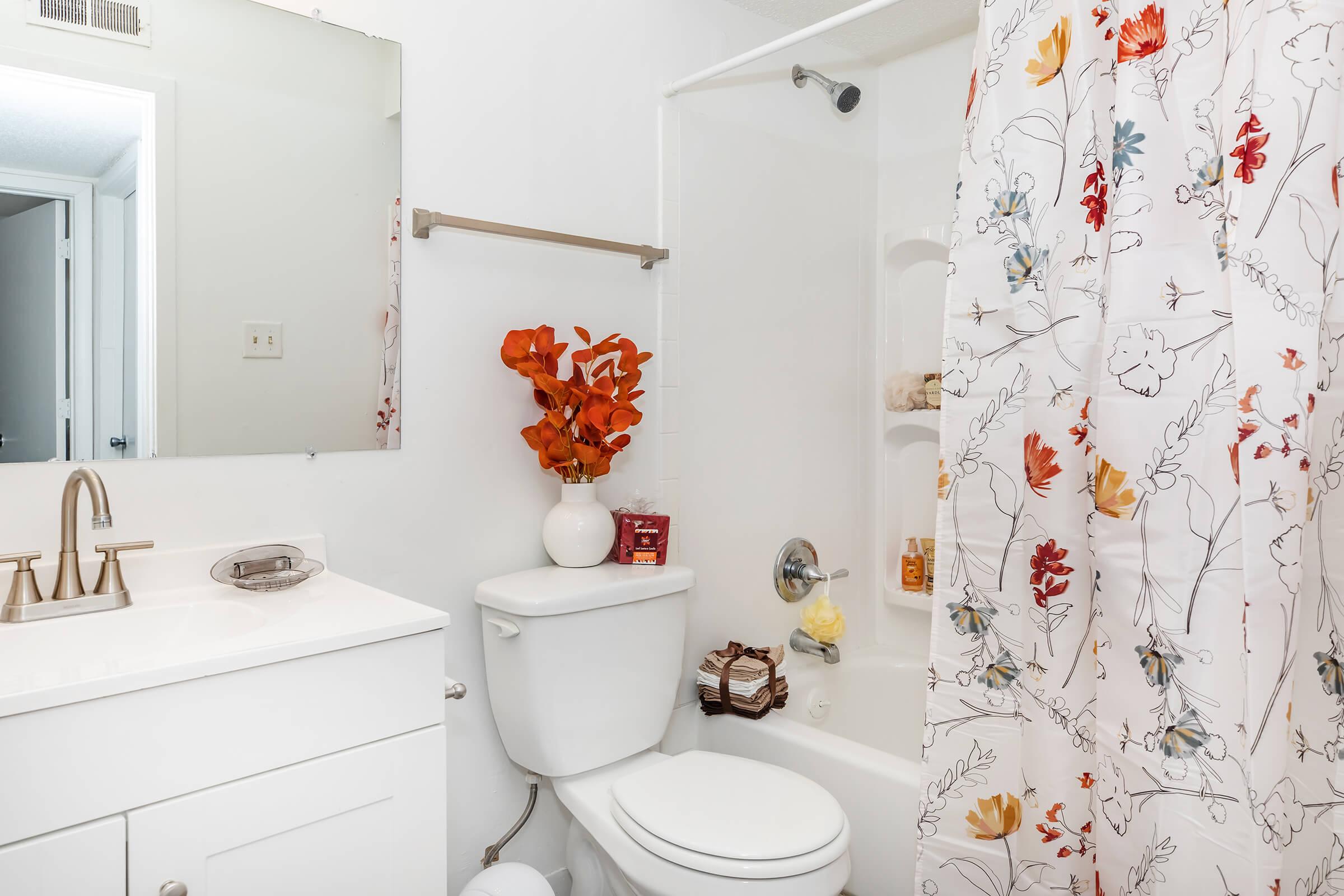 A bright bathroom featuring a white vanity with a sink and mirror, a toilet, and a bathtub with a floral shower curtain. The bathtub holds various toiletries, and a vase with orange flowers sits on the vanity. The walls are painted white, and there's a glimpse of a door leading to another room.