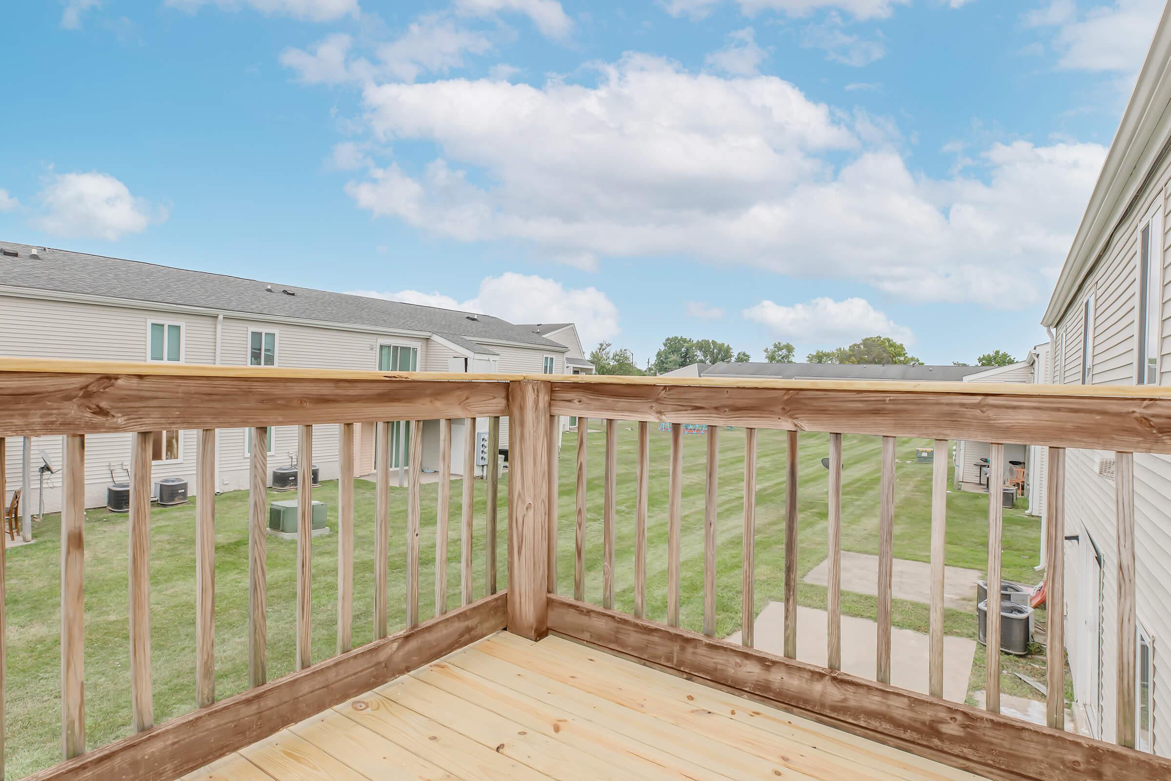 View from a balcony showcasing a wooden railing overlooking a grassy yard. In the distance, there are two-story buildings with several air conditioning units. The sky is partly cloudy, creating a serene outdoor atmosphere.