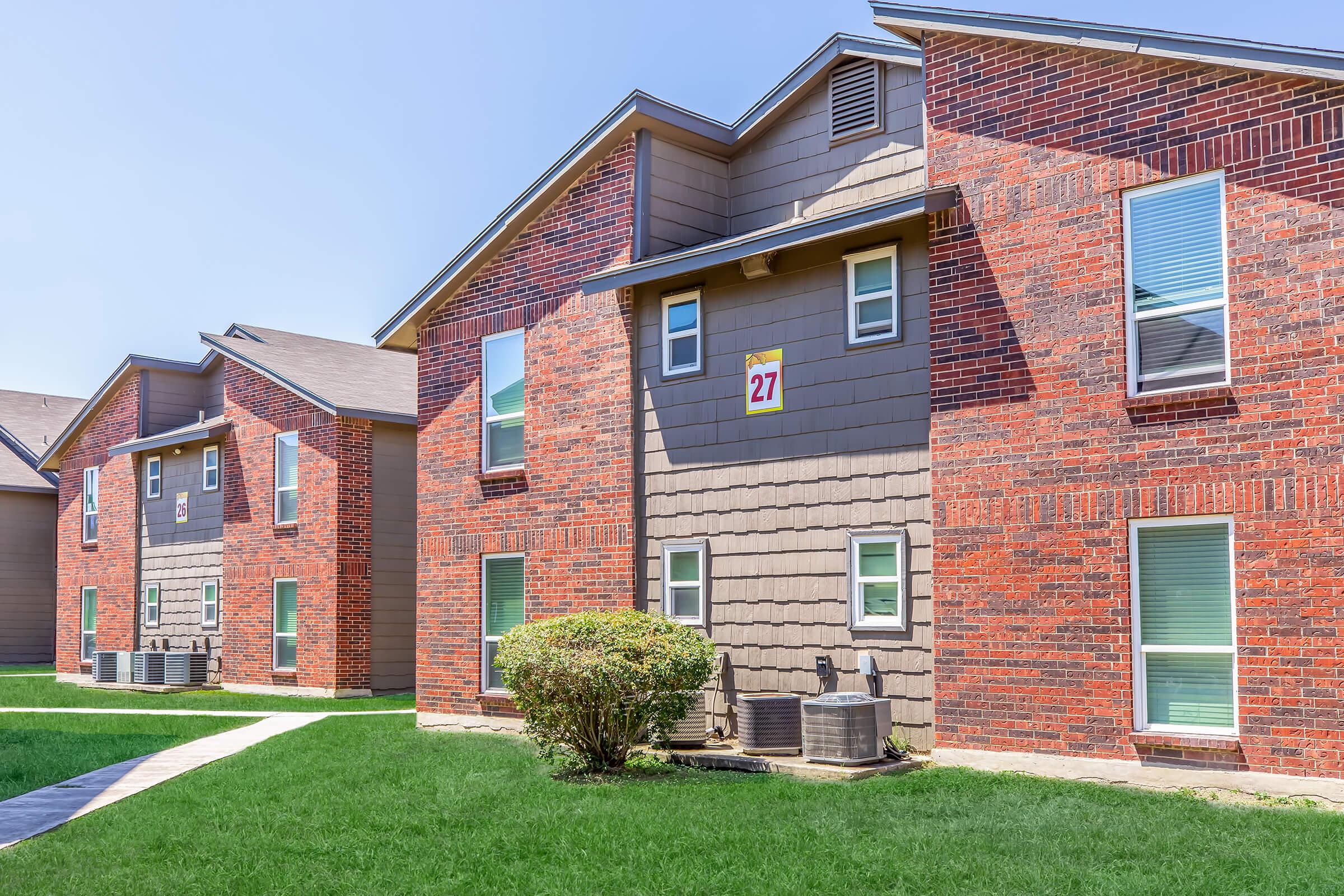 Two multi-unit apartment buildings with a mix of brick and siding exteriors. The building on the right features a large window and a number "27" displayed above the entrance. Lush green grass surrounds the buildings, with a paved walkway leading between them under a clear blue sky.