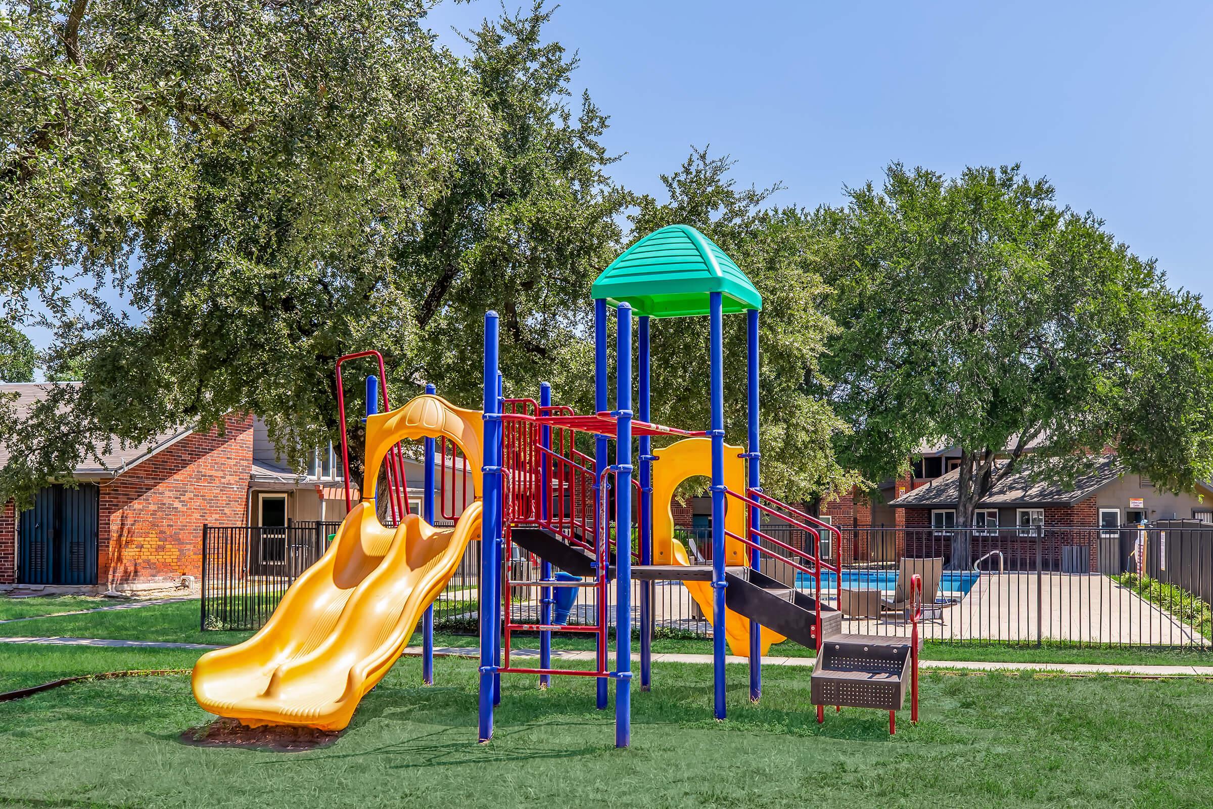 A colorful playground featuring a yellow slide, climbing structure, and green roof, set in a grassy area with trees. A pool is visible in the background, surrounded by a black fence. The playground is designed for children, providing a fun and safe environment for play.