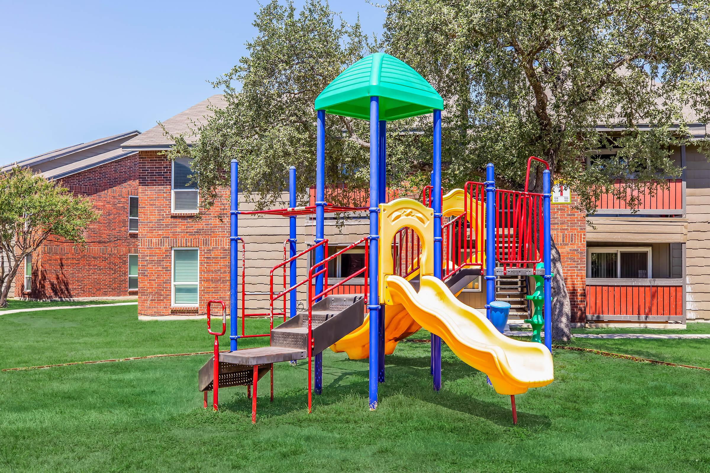 Colorful playground equipment with a green roof, yellow slide, and multiple climbing structures set on a grassy area. Surrounding the playground are residential buildings with brick and stucco exteriors, under a clear blue sky.