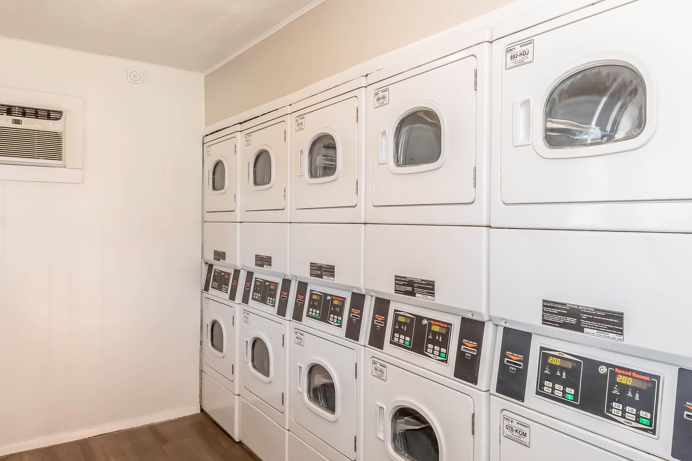 A clean and organized laundry room featuring several stacked washing machines and dryers. The machines have digital control panels with various settings, and the walls are painted a light color, creating a bright environment. An air conditioning unit is visible on the left side.