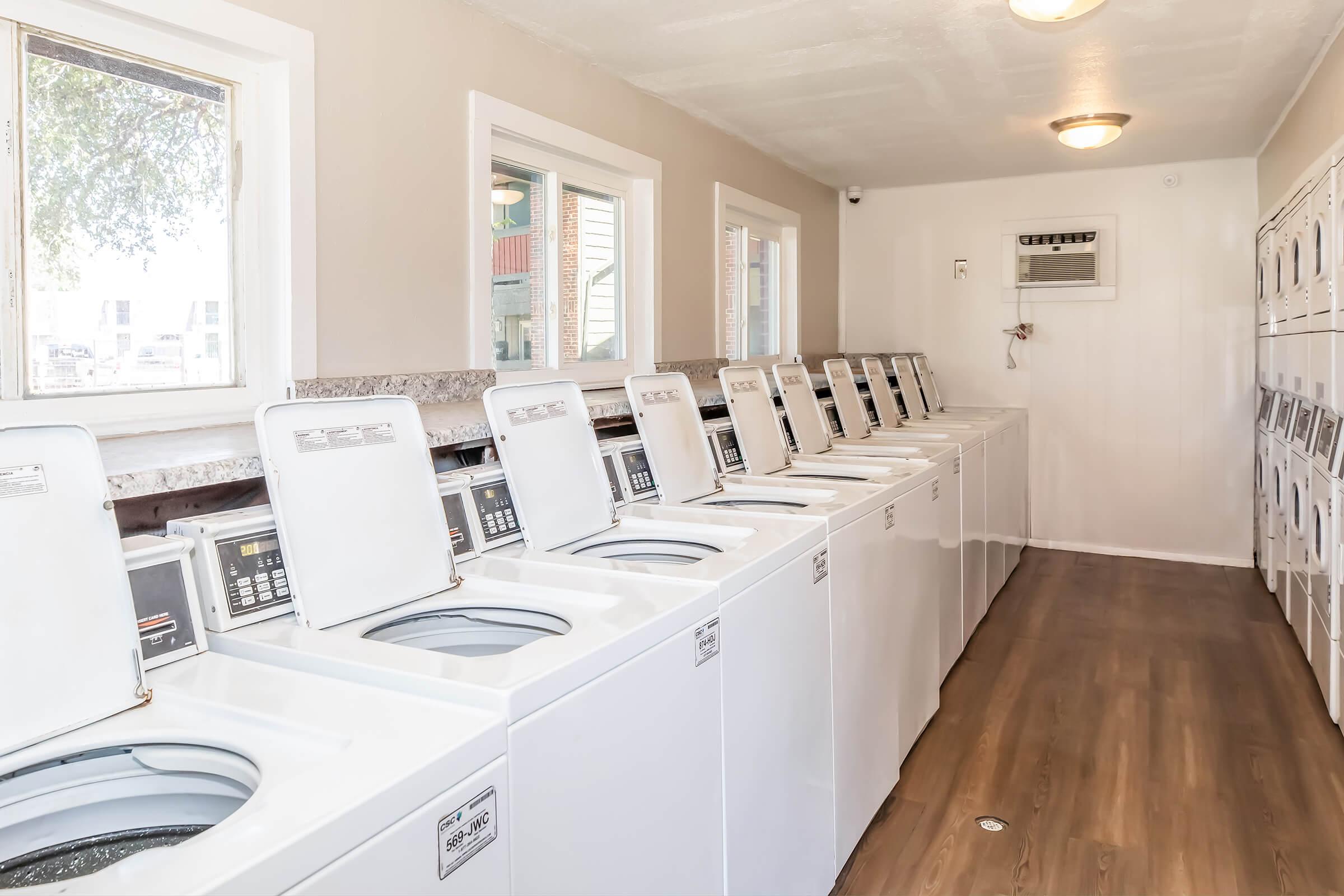 A clean and well-lit laundry room featuring a row of white washing machines and dryers. The space has windows that provide natural light, with polished wooden flooring and a neutral-colored wall. The machines are neatly arranged, creating an organized and functional environment for doing laundry.