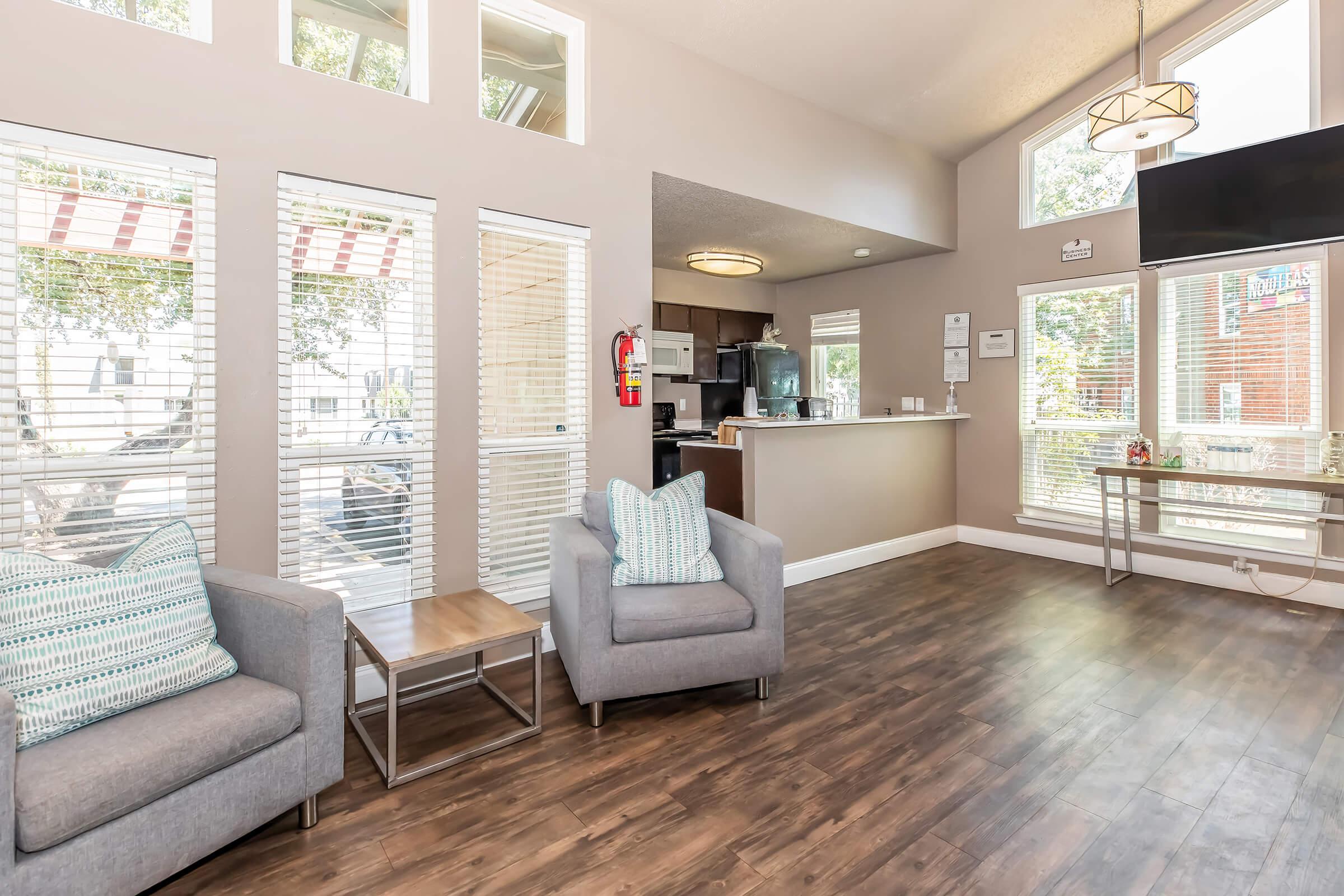 Bright and modern common area featuring two grey armchairs with decorative pillows, a small wooden side table, and large windows allowing natural light. In the background, a kitchenette and a reception area are visible, with a stylish chandelier overhead and a TV on the wall.