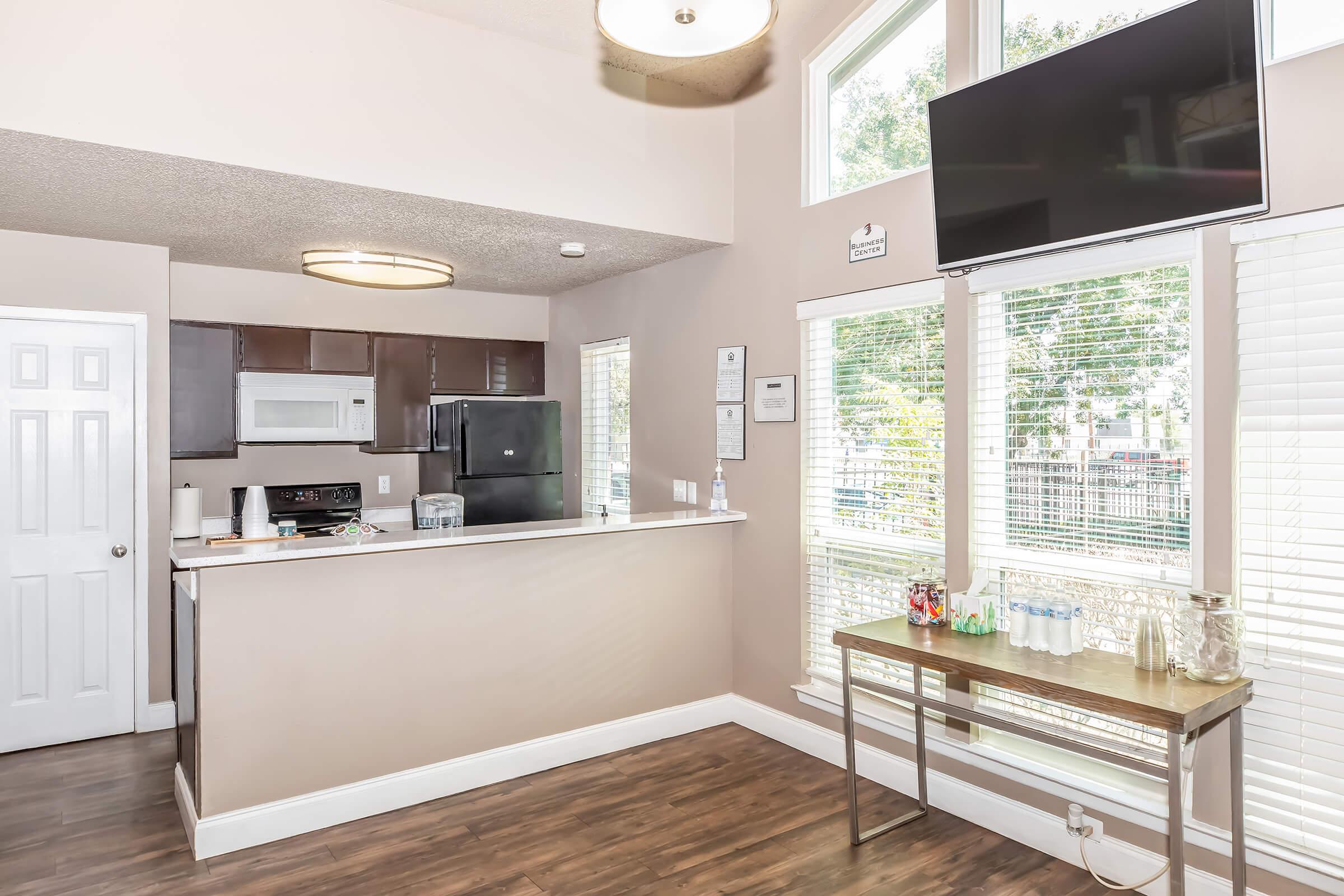 A modern kitchen area with a bar counter, featuring a microwave, refrigerator, and a television mounted on the wall. Large windows provide natural light, with a view of an outdoor area. A table displays bottled water and a glass jar. The space has light-colored walls and wood-style flooring.