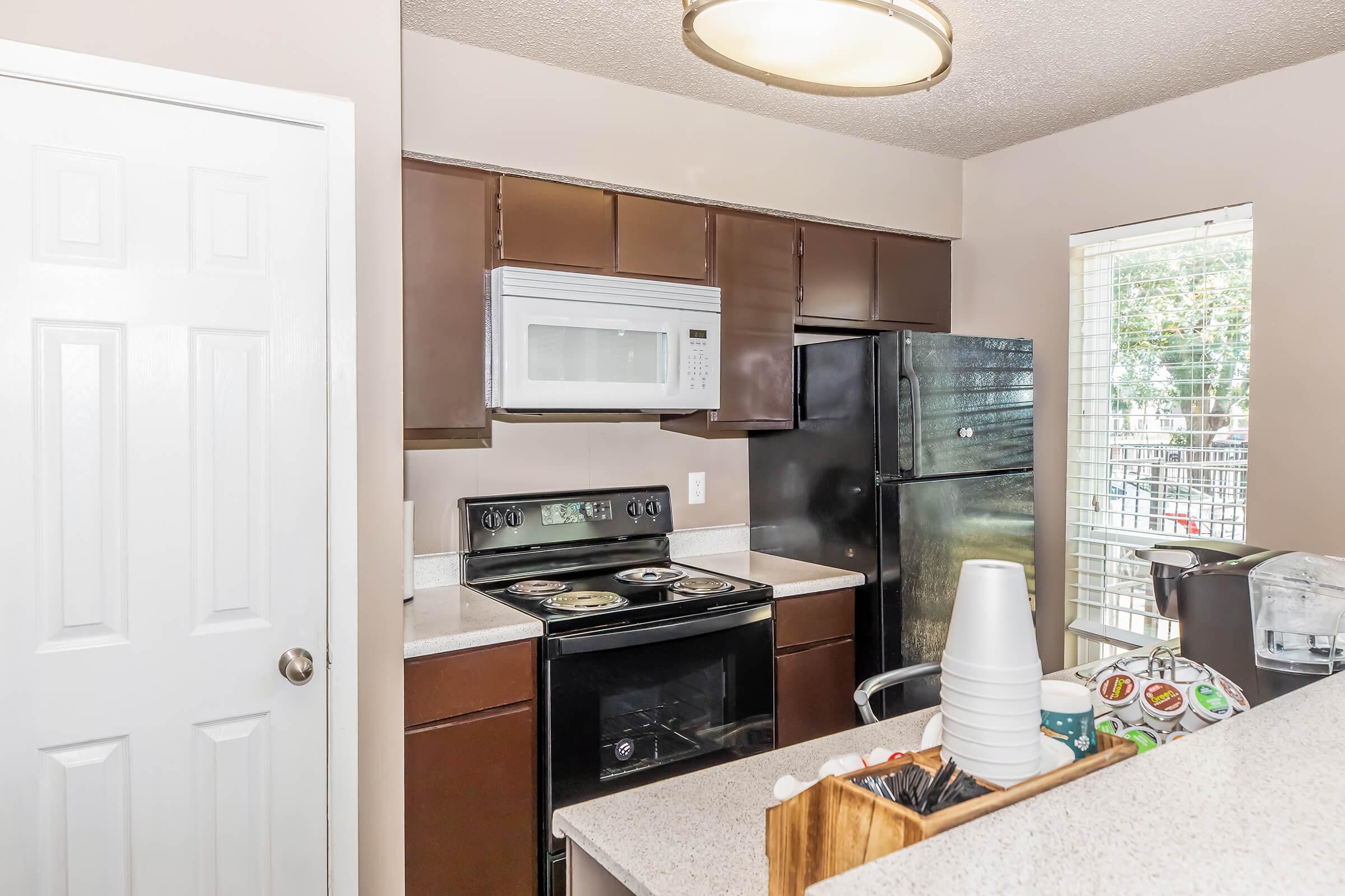 A modern kitchen featuring dark wooden cabinets, a stainless steel microwave above a black stove and oven, and a black refrigerator. A light fixture hangs above, and a granite countertop holds various kitchen items. A window allows natural light, revealing a view outside.