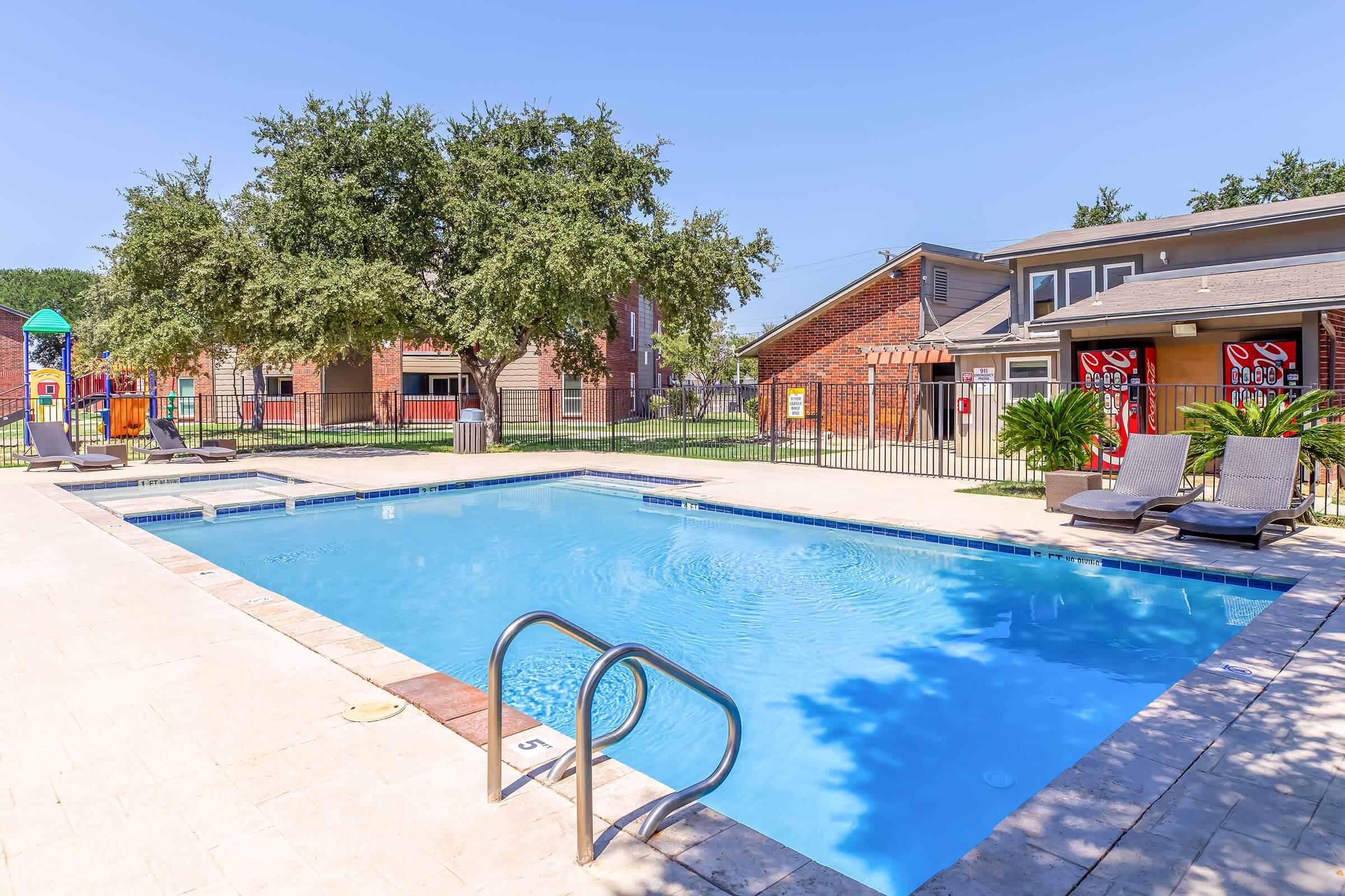 A clear blue swimming pool surrounded by a patio area, featuring two lounge chairs on one side. In the background, there are trees and residential buildings, with a children’s play area visible. The scene is bright and sunny, indicating a pleasant outdoor environment.