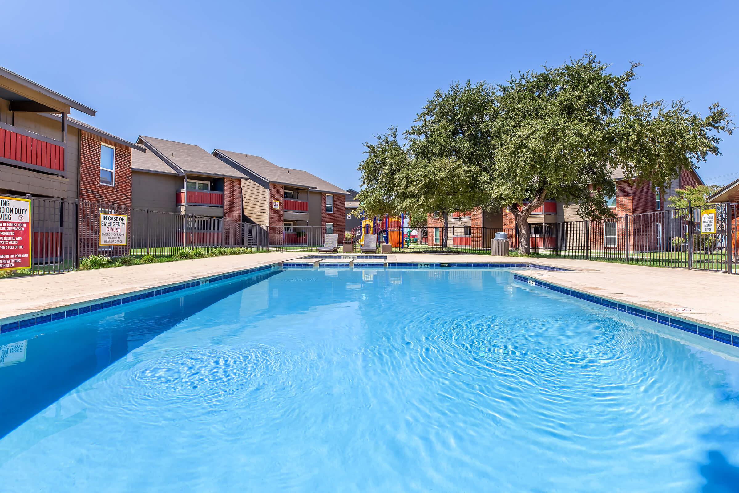 A clear blue swimming pool surrounded by a sunny courtyard. In the background, there are two-story apartment buildings with red accents and a large oak tree providing shade. The scene conveys a refreshing and inviting atmosphere perfect for relaxation and leisure activities.