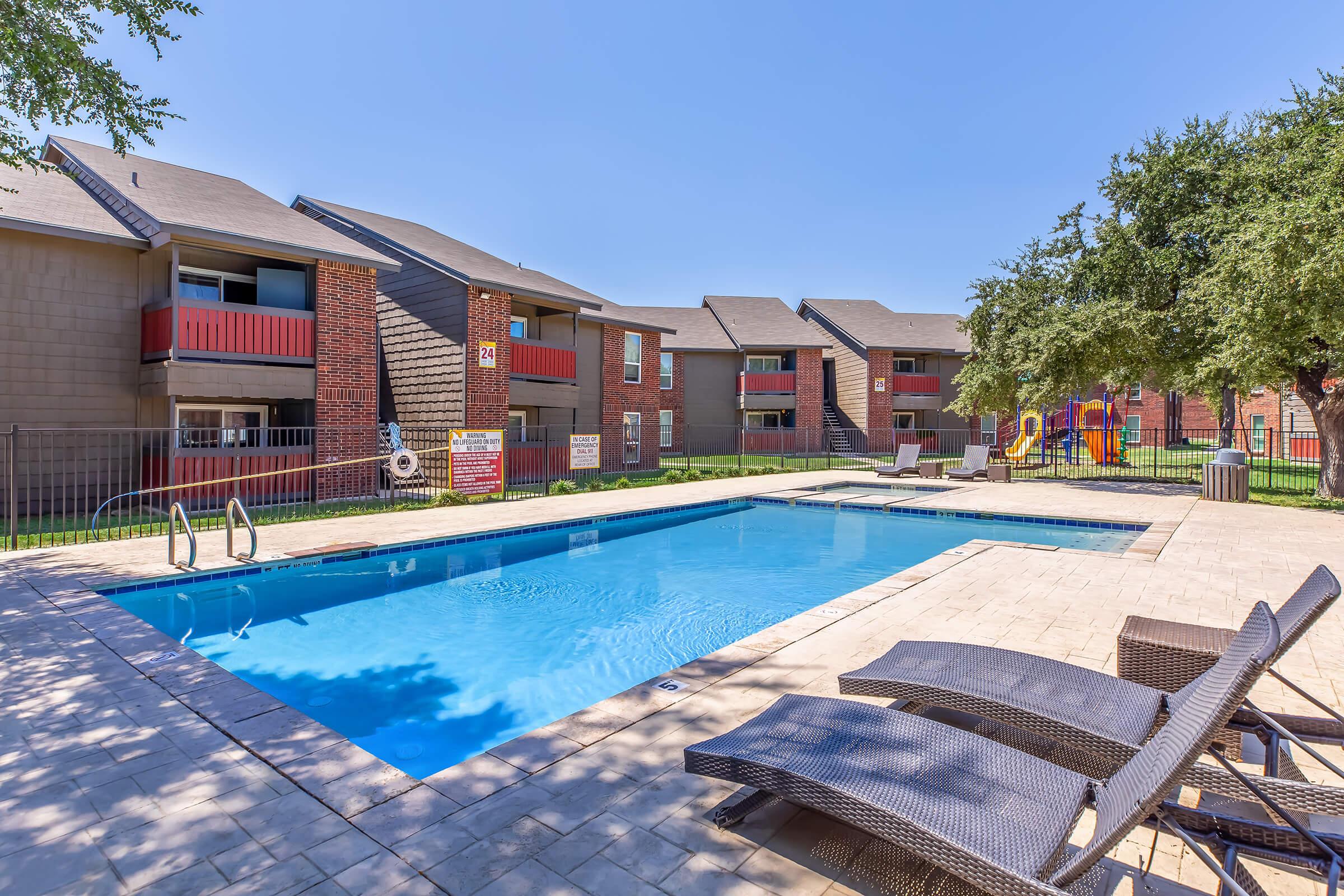 A clear blue swimming pool surrounded by lounge chairs and a fence, with several apartment buildings in the background. The scene features green lawns and trees, creating a relaxing outdoor space. A playground is visible nearby, adding to the family-friendly atmosphere.