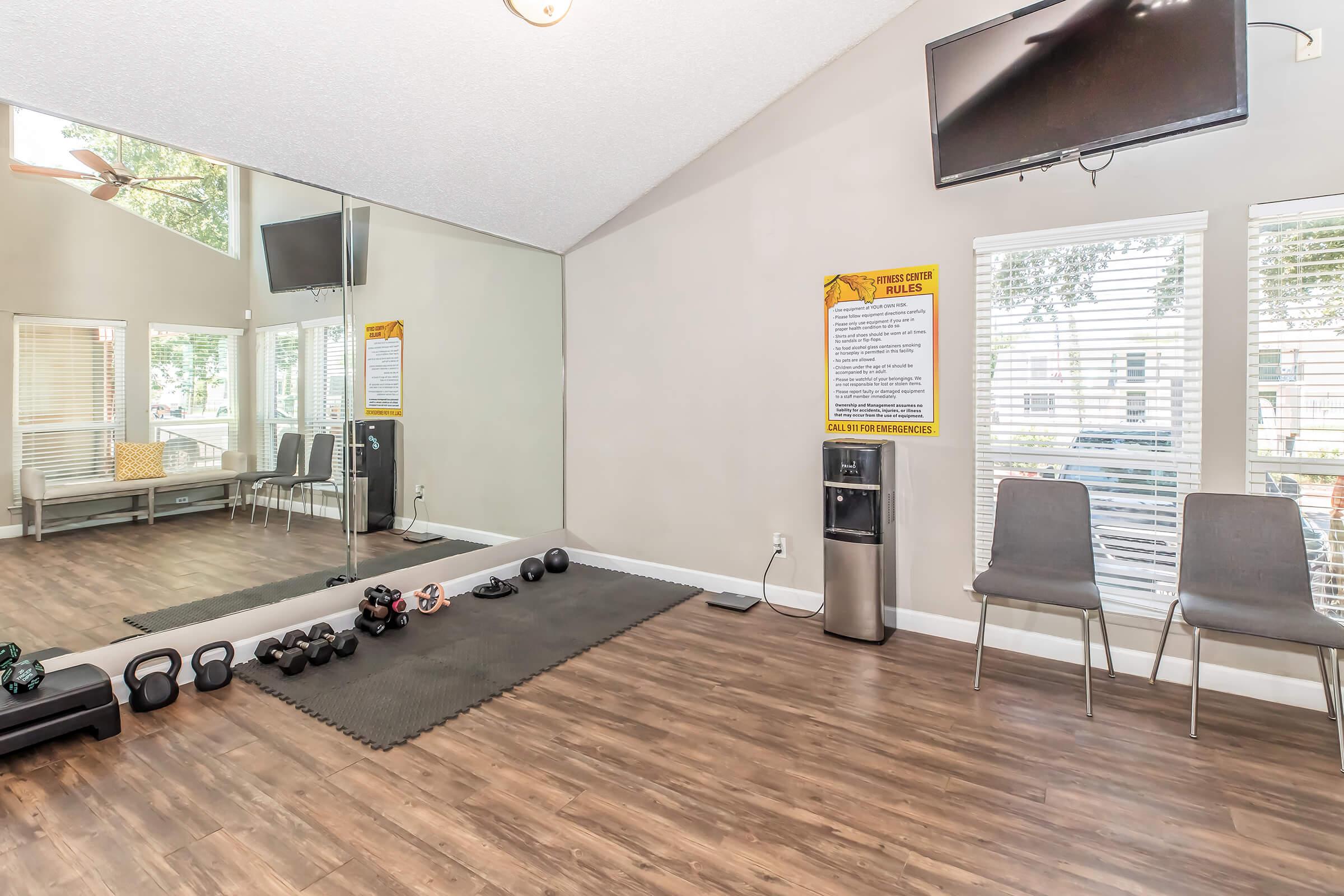 A well-lit gym area featuring a mirrored wall, exercise mats, and a variety of dumbbells. There is a water cooler and two chairs beside large windows with blinds. A television is mounted on the wall, and a fitness instructions poster is visible. The space is designed for workouts and fitness activities.
