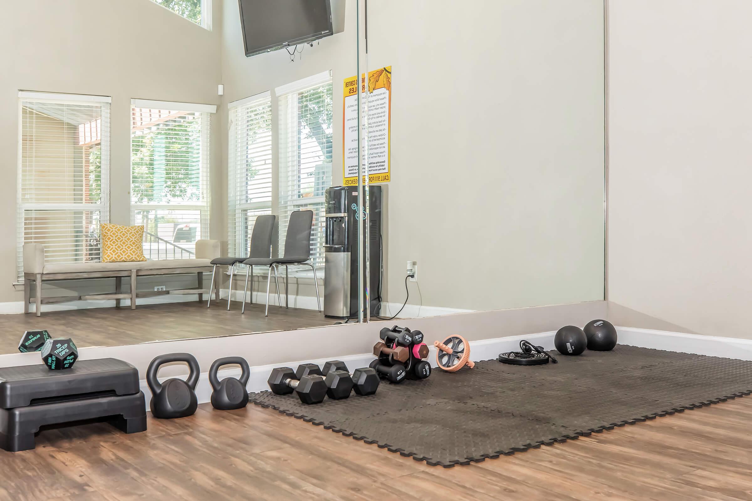 A fitness space featuring a rubber mat on the floor with various weights, including dumbbells, kettlebells, and a barbell. In the background, there are large windows with natural light, a seating area with two chairs and a cushion, and a television mounted on the wall.