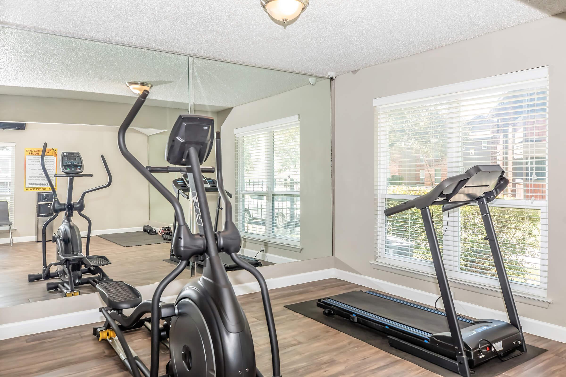 A fitness room featuring an elliptical machine and a treadmill, with a large wall mirror reflecting the equipment. Natural light filters through a window with blinds, and the floor is made of wooden planks, creating a modern and inviting workout space.