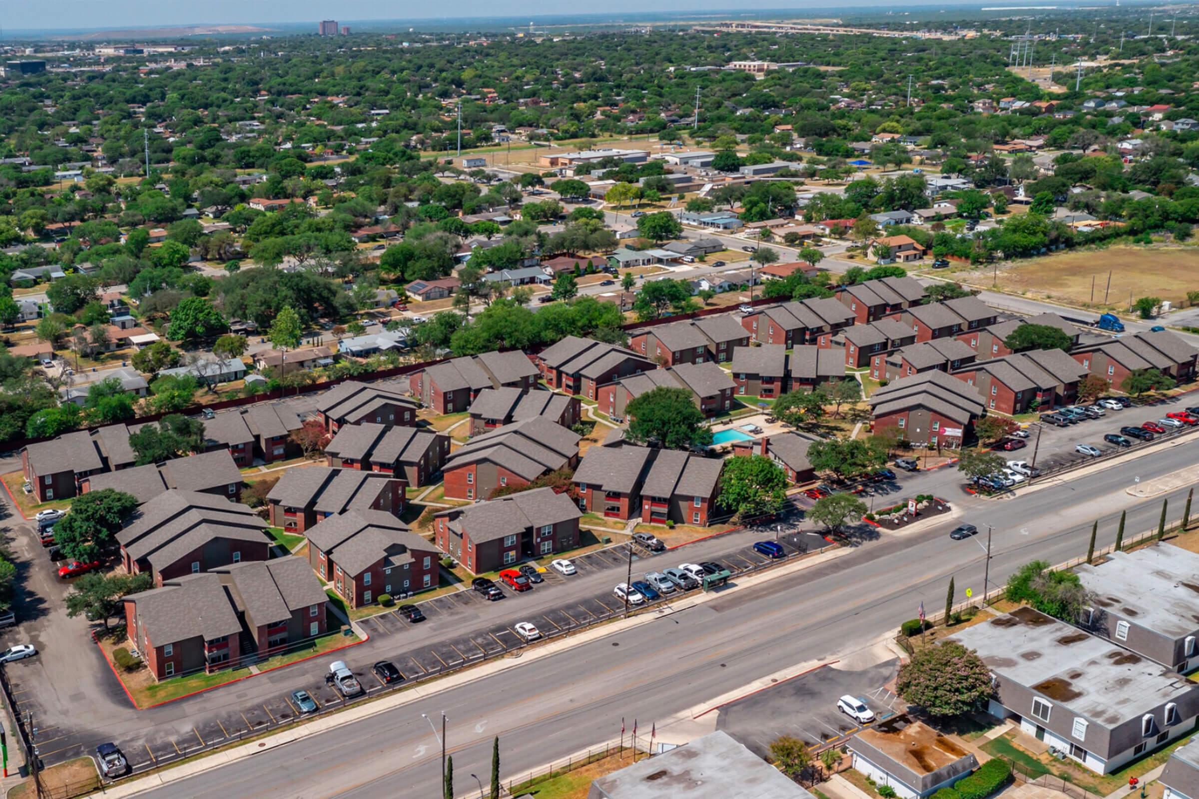 Aerial view of a residential neighborhood featuring several two-story apartment buildings arranged around a central pool area. Surrounding greenery and a road with parked cars can be seen, along with other homes in the distance. The scene captures a suburban landscape with a mix of housing and greenery.