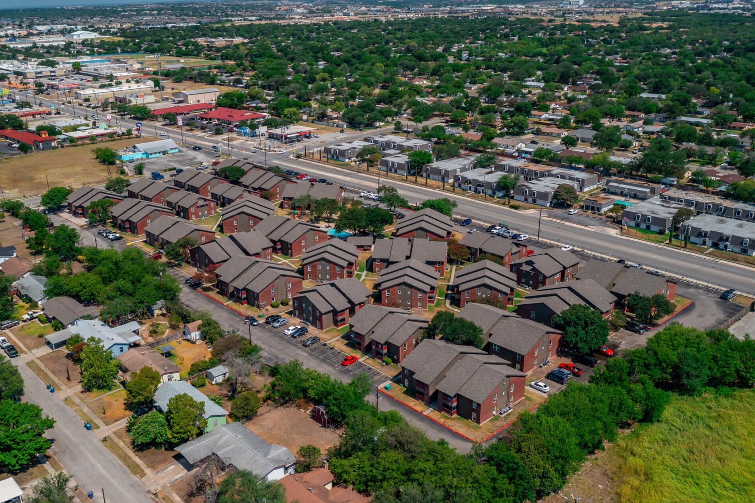 Aerial view of a residential area featuring multiple buildings, likely apartments or townhouses, surrounded by greenery and parking lots. Nearby, a main road runs alongside the development, with various trees and a mix of residential and commercial structures in the background.