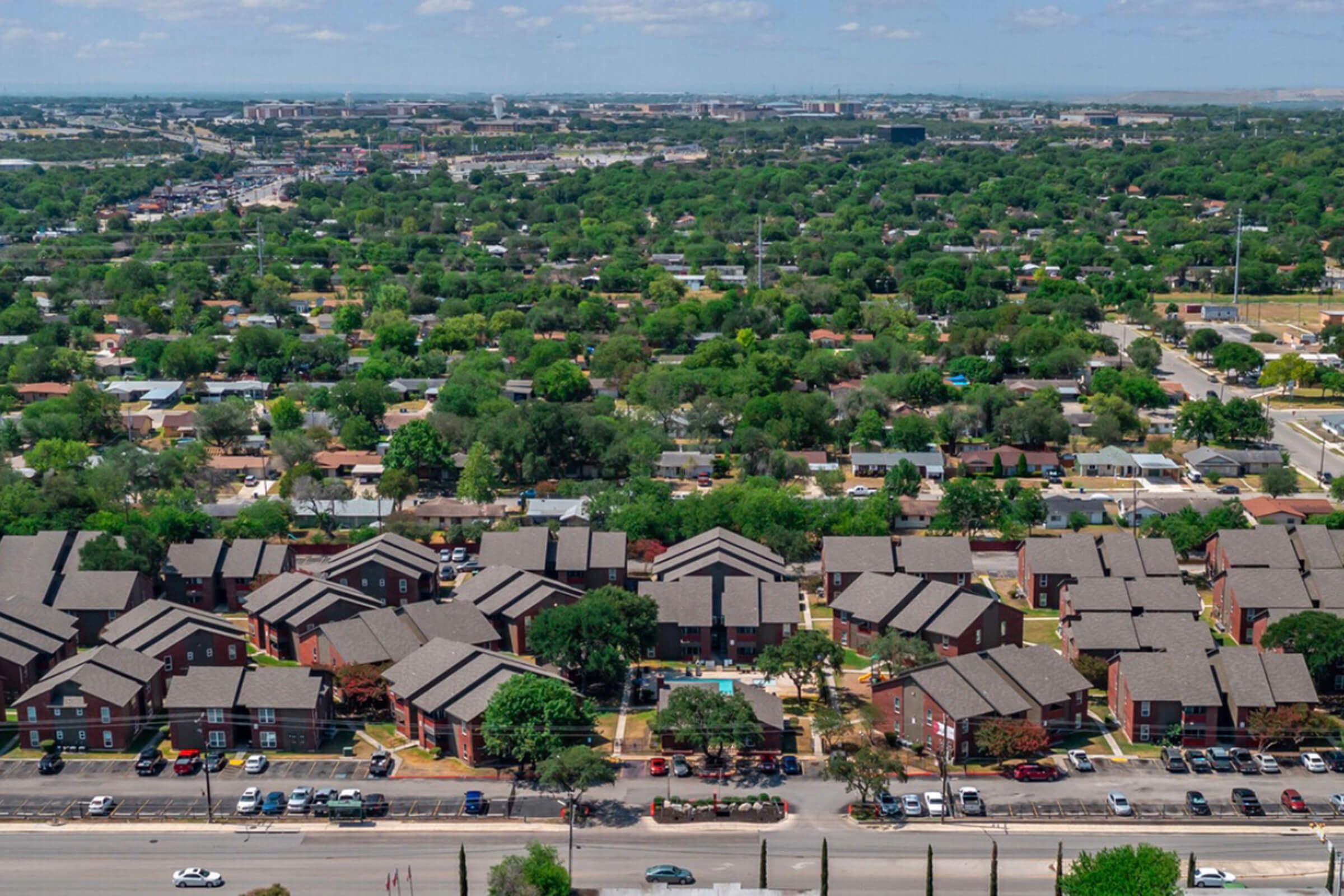 Aerial view of a residential neighborhood featuring multiple low-rise apartment buildings, surrounded by lush greenery and trees. The landscape includes a mix of houses and roads, with vehicles parked along the streets. Distant buildings and urban infrastructure are visible in the background under a clear blue sky.