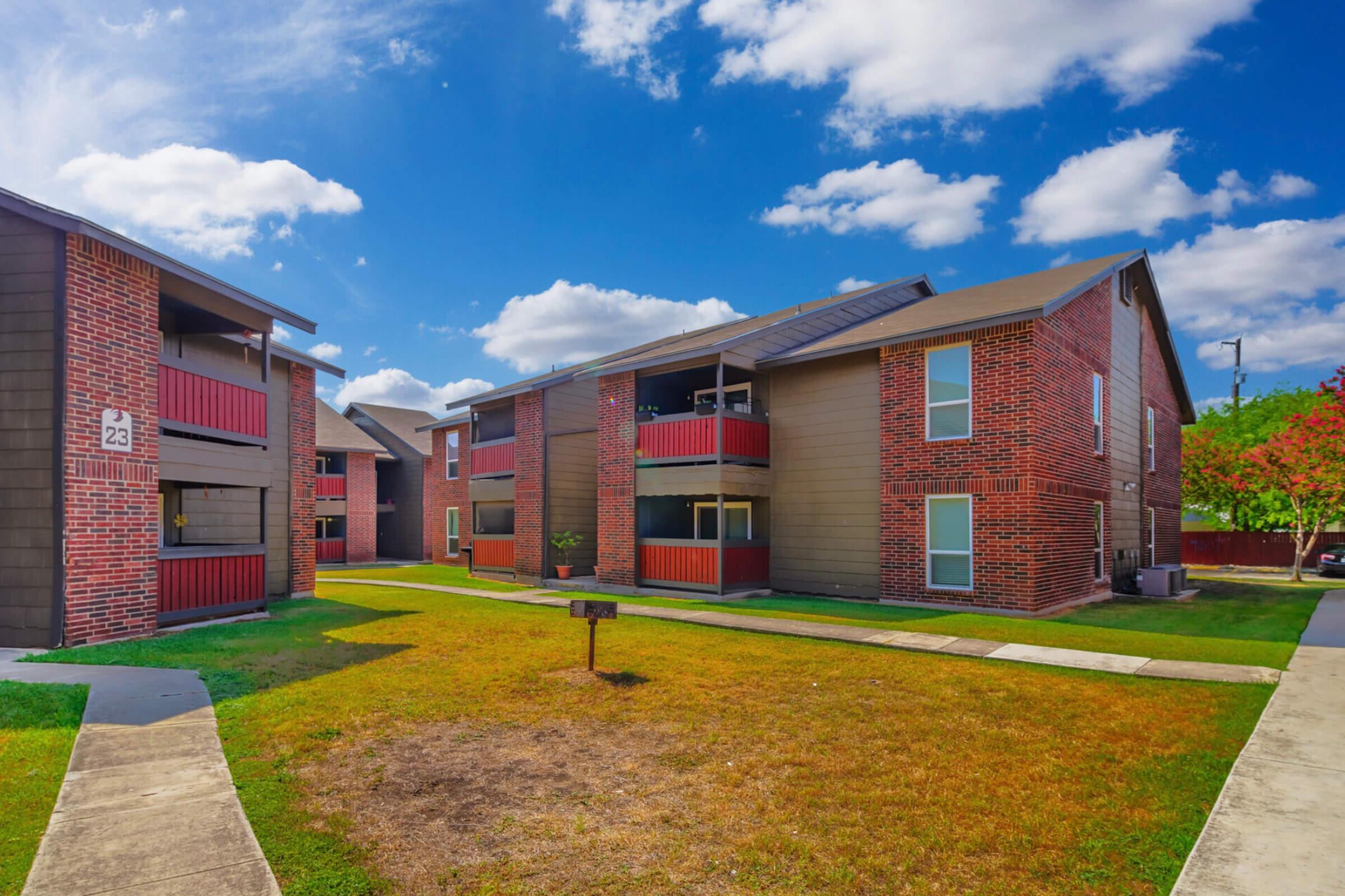View of a residential apartment complex featuring two buildings with red brick exteriors and balconies. The foreground shows a grassy area with a sidewalk and a small barbecue grill. The sky is bright blue with scattered clouds, and the scene is well-lit, suggesting a sunny day.