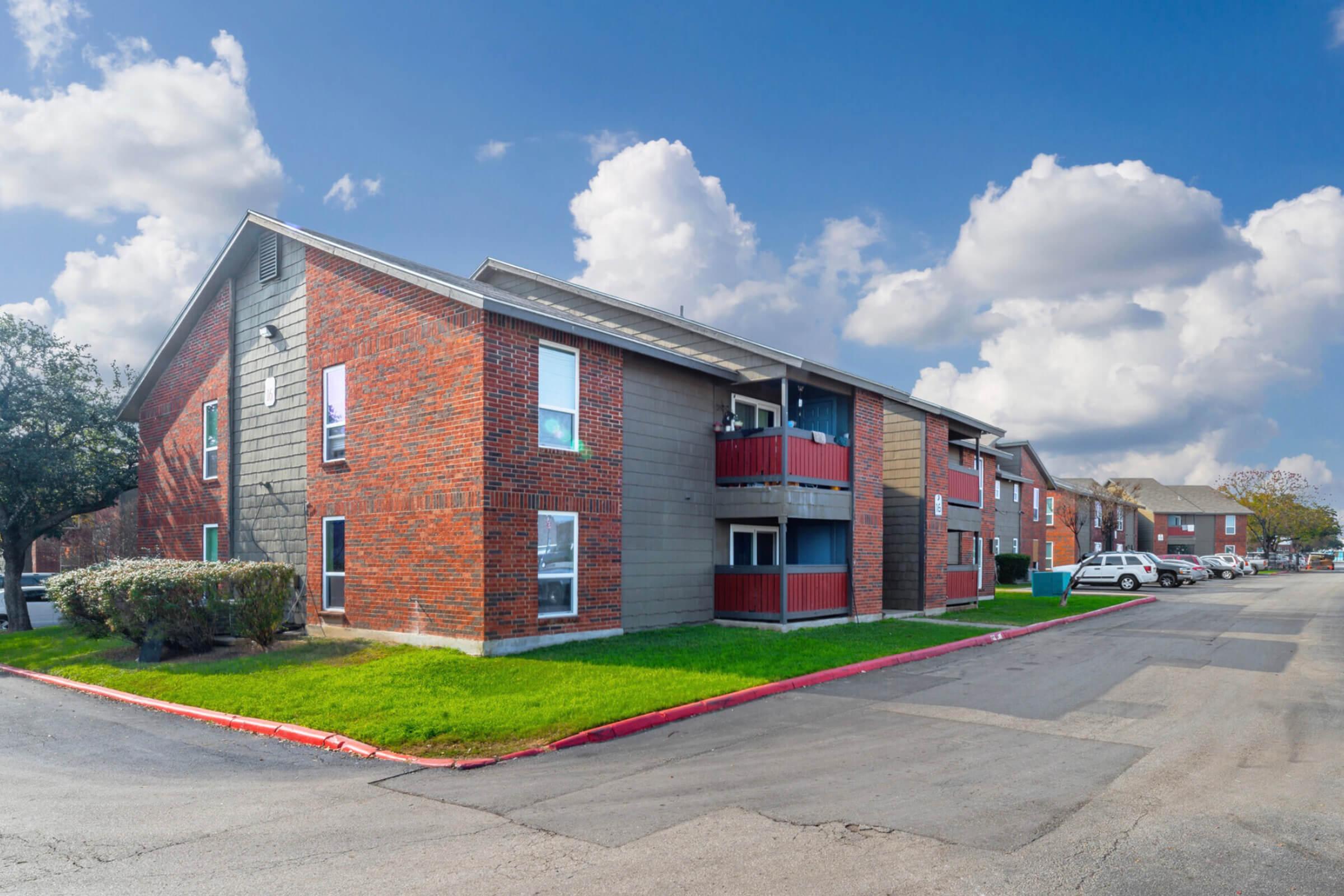 A three-story brick apartment building with a mix of red and gray siding. The building features several windows and balconies. A landscaped area with grass and small bushes is in front. A paved parking lot runs alongside the building, with a few parked cars visible. The sky is partly cloudy.