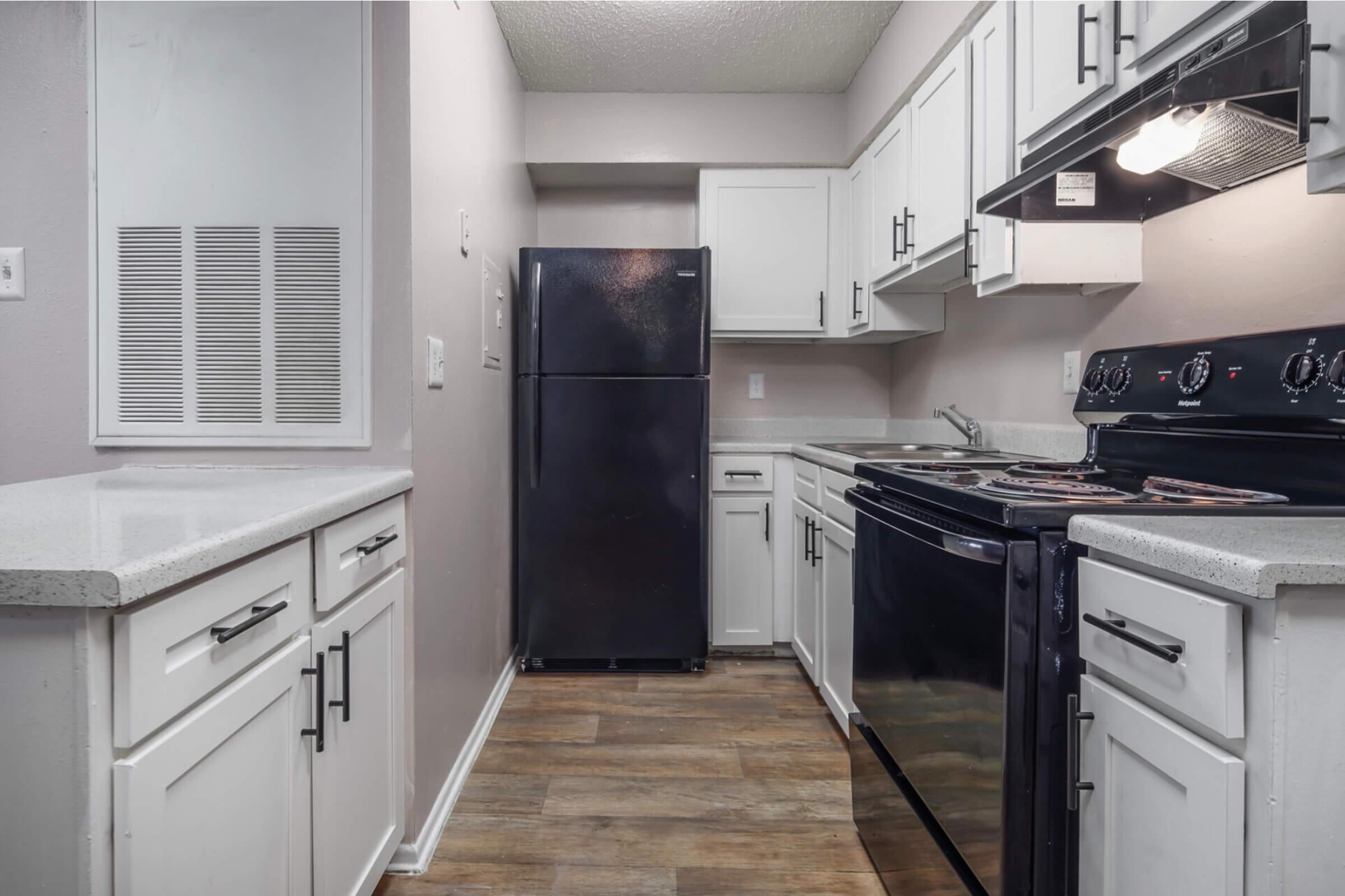 A modern kitchen featuring white cabinets, a black refrigerator, a black stove with an oven, and a white countertop. The walls are painted in a light shade, and there's a vent on the left side. The overall design is clean and contemporary.