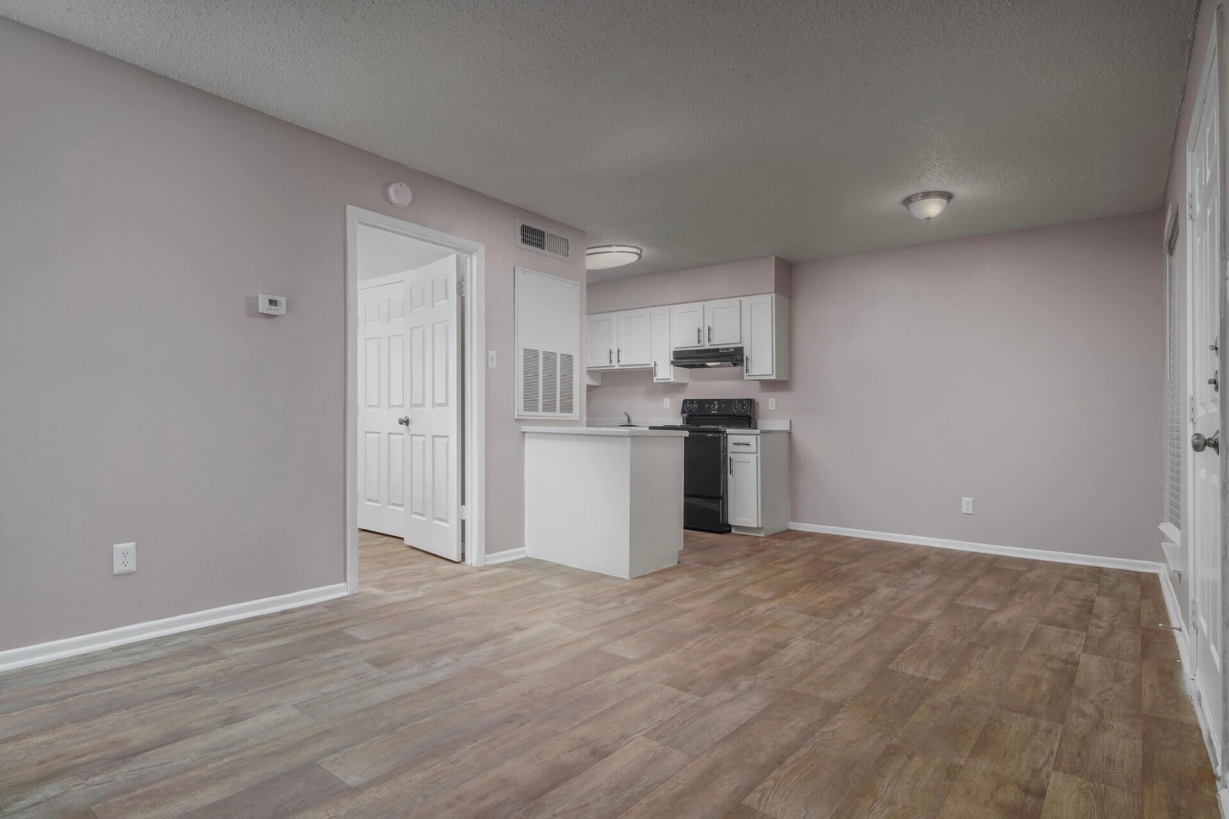 A modern, unfurnished living space featuring light pink walls, vinyl flooring, and a kitchen area with white cabinets and black appliances. A partition separates the kitchen from the main room, and there are double doors leading to a closet. Natural light fills the area, creating a bright and inviting atmosphere.