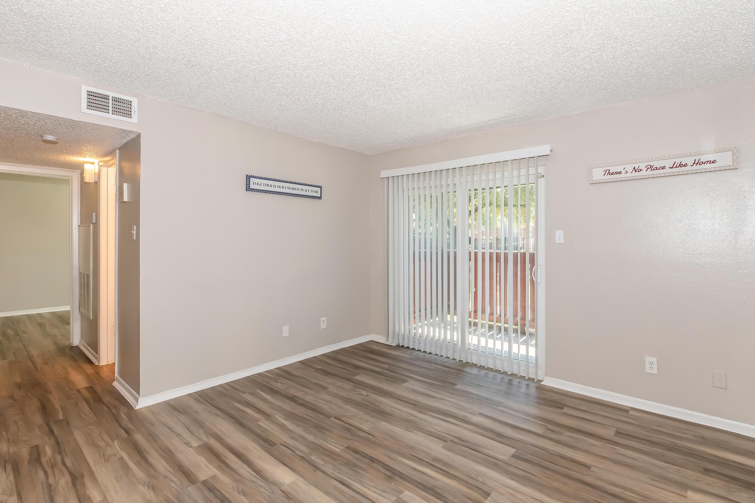 A spacious living room with light-colored walls and wooden flooring. It features a large sliding glass door with vertical blinds leading to an outdoor area. A small wall plaque reads, "There's No Place Like Home." A hallway is visible in the background, leading to another room.