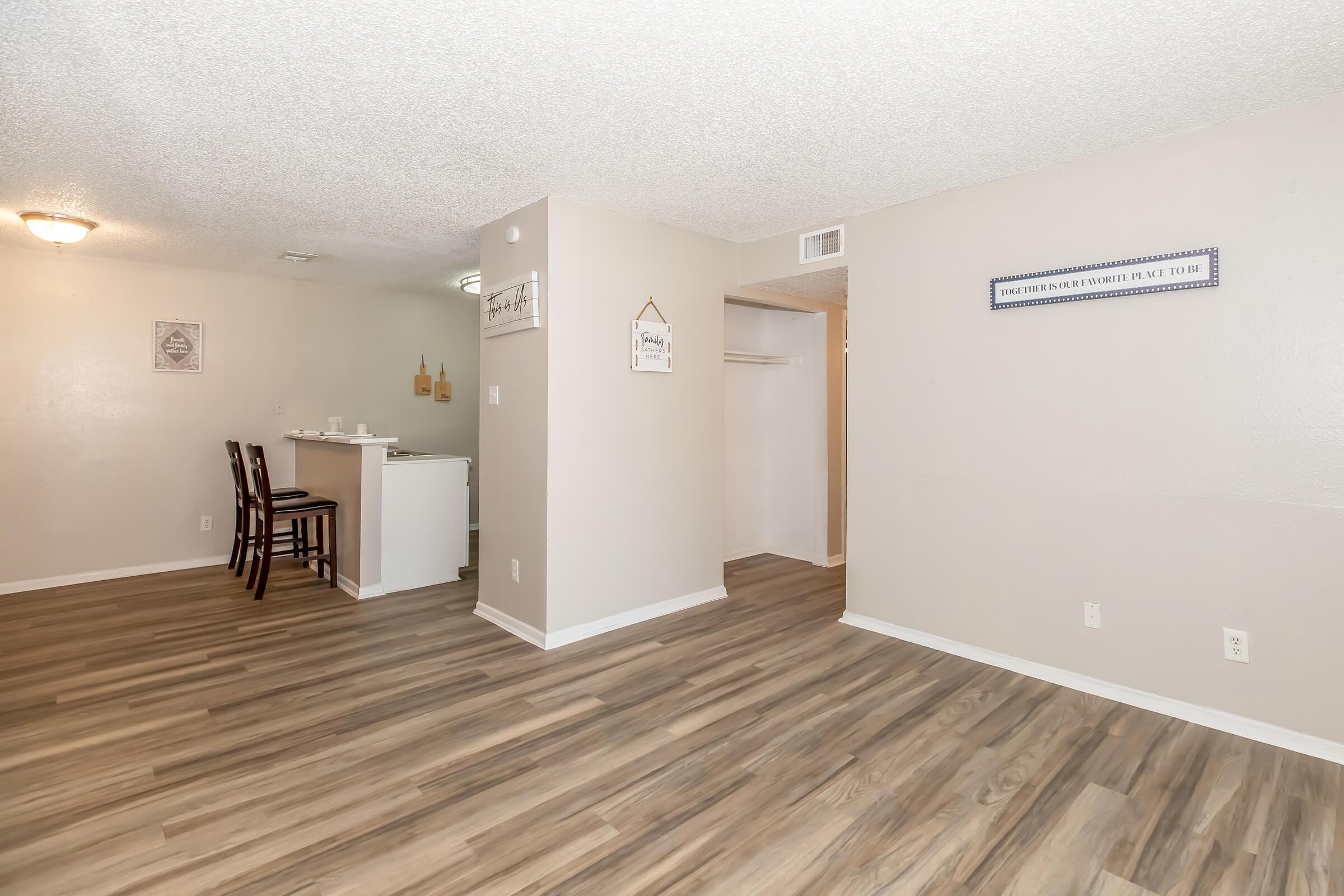 Interior view of a modern apartment featuring an open layout with hardwood flooring. On the left, a dining area with a small table and two chairs. A kitchen counter is visible, and a closet space can be seen in the background. Walls are painted in neutral tones, creating a bright and spacious atmosphere.