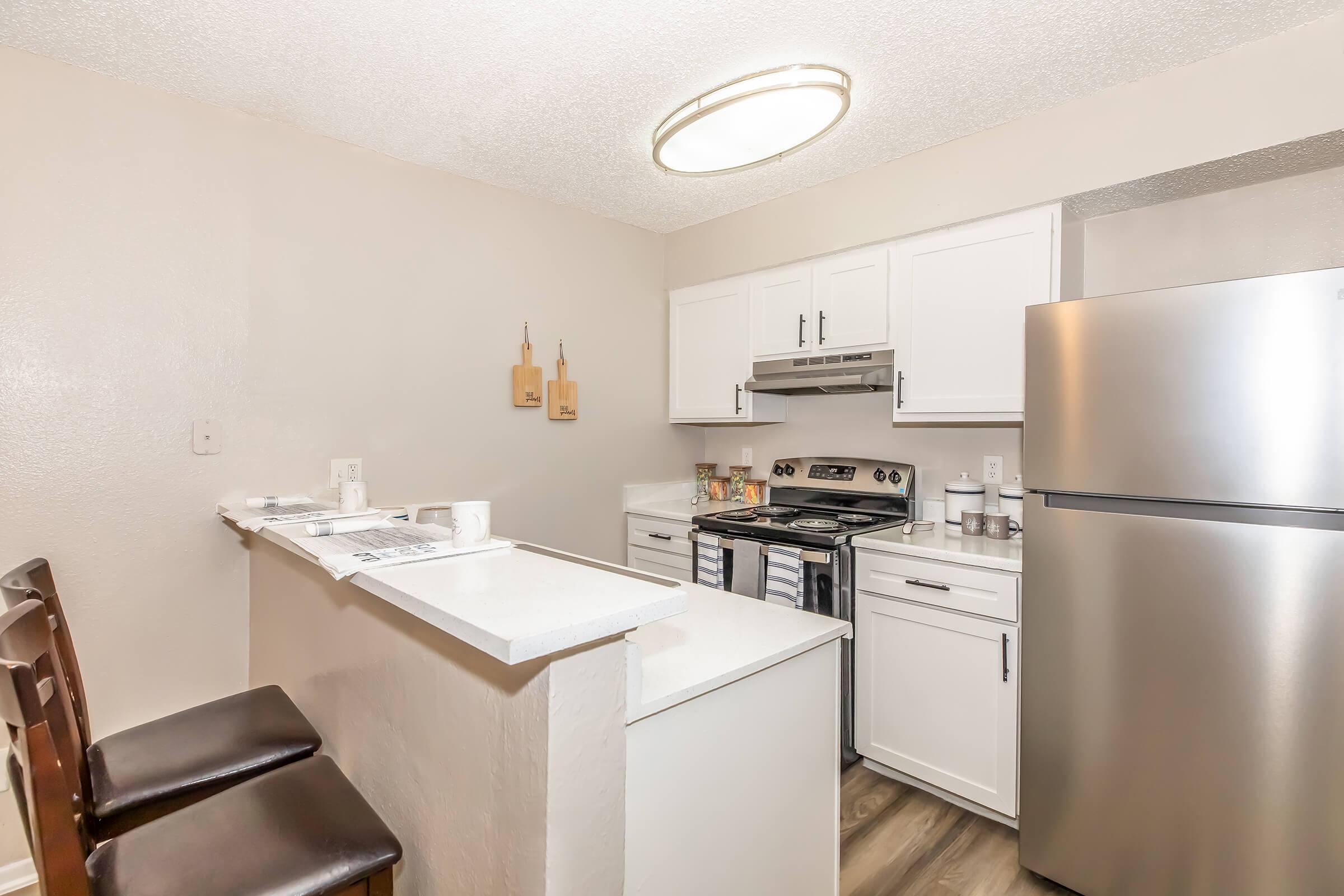 A modern kitchen featuring white cabinetry, stainless steel appliances including a refrigerator and stove, a white countertop, and two brown bar stools at the kitchen island. The walls are painted a light color, and there are wooden decor items on display.