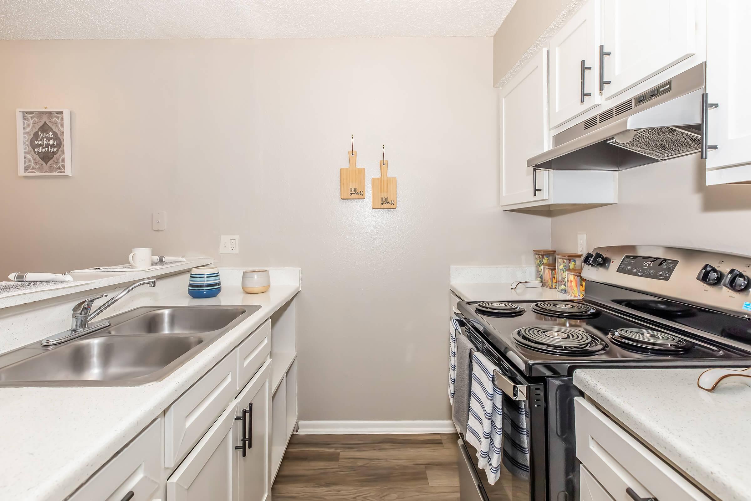 A modern kitchen featuring white cabinetry, a double sink, and a black stove. Two wooden cutting boards are mounted on the wall. The countertop is decorated with dishware and kitchen accessories. The flooring is a warm, wood-like laminate.