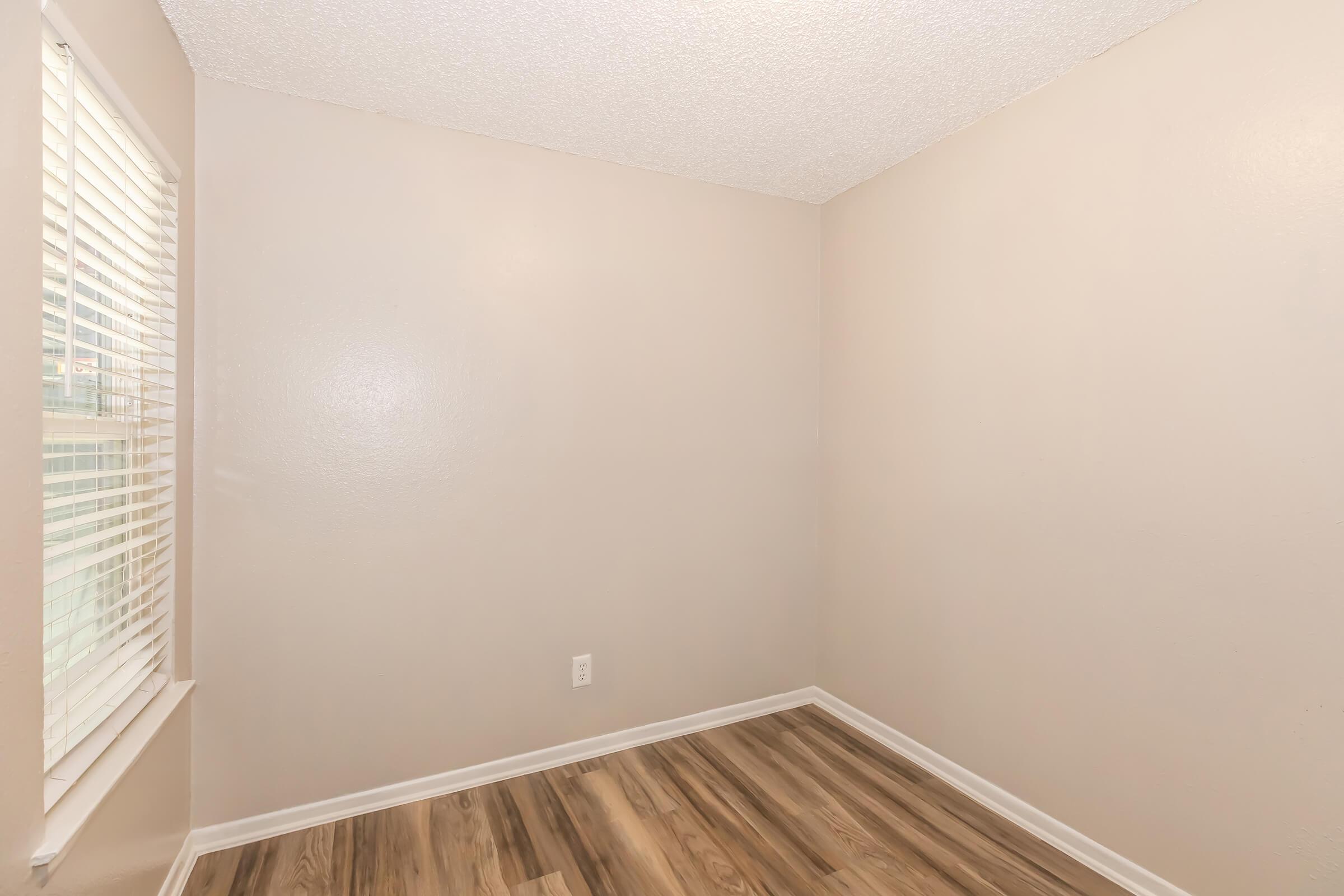 A small, empty room with light beige walls and a textured ceiling. The floor is made of light wood-like laminate. There is a window with white blinds allowing natural light to enter the space. The room has no furniture, creating a clean and open atmosphere.