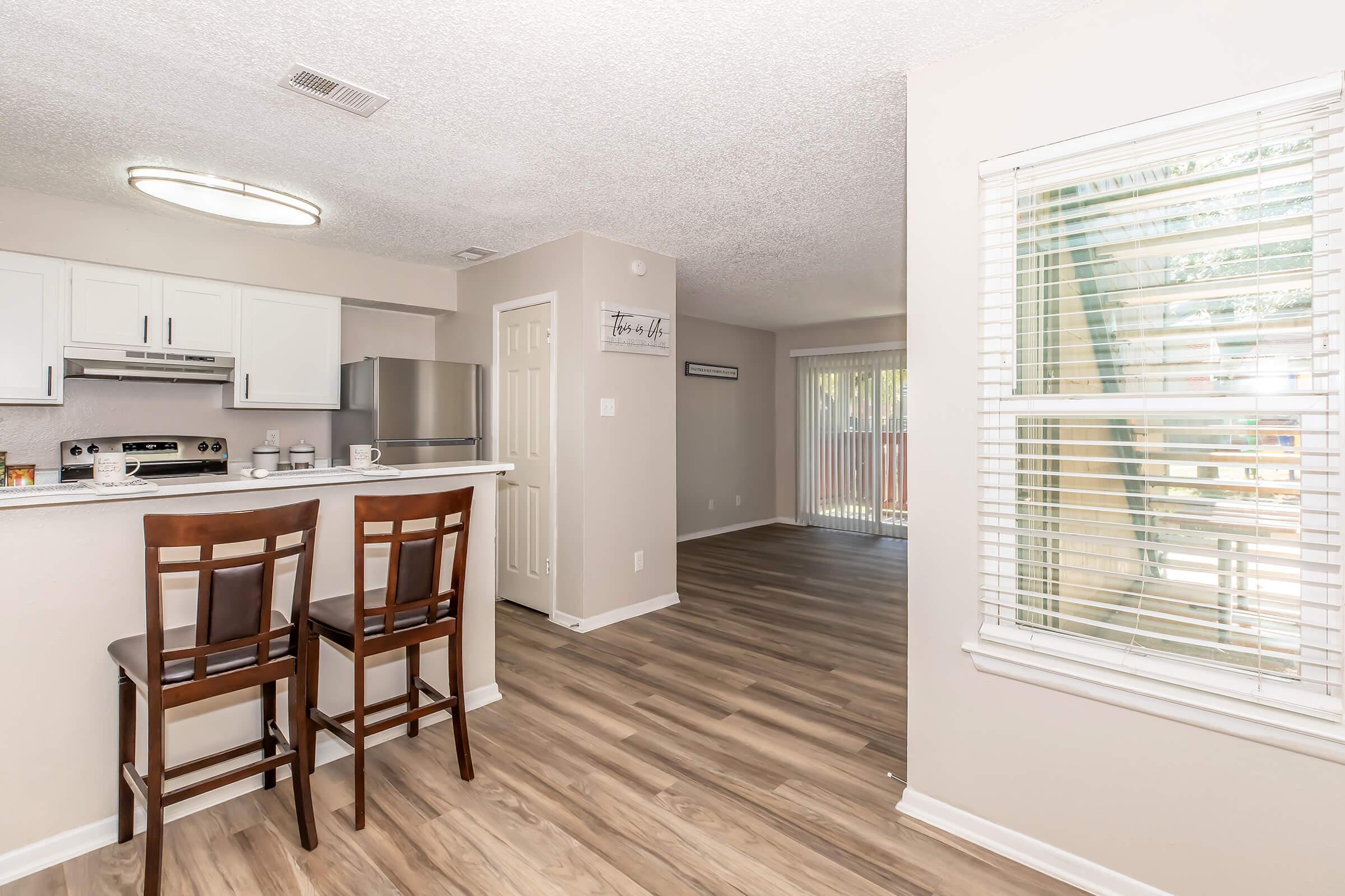 Modern open-concept kitchen and living area featuring a breakfast bar with two stools, sleek white cabinetry, stainless steel appliances, and large windows allowing natural light. The living area has sliding glass doors leading to an outdoor space, with light-colored flooring throughout.