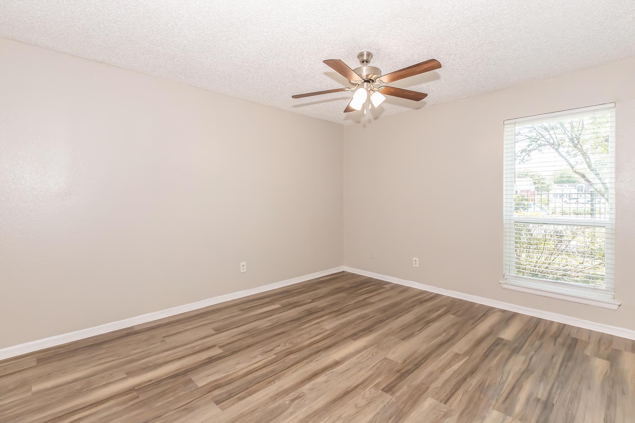 A spacious, empty room featuring light beige walls, wood-style flooring, and a ceiling fan with wooden blades. A window allows natural light to enter, providing a view of the outdoors. The room has no furniture, creating a clean, open space ideal for various uses.