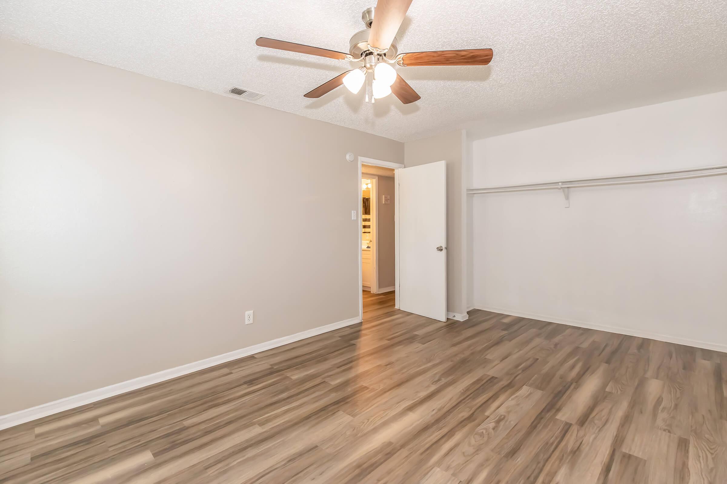 A spacious room with light-colored walls and a ceiling fan featuring wooden blades. The floor is made of laminate with a wood-like finish. A doorway leads to another area, and there is a closet with a shelf along one wall, providing ample storage space. The overall ambiance is bright and inviting.