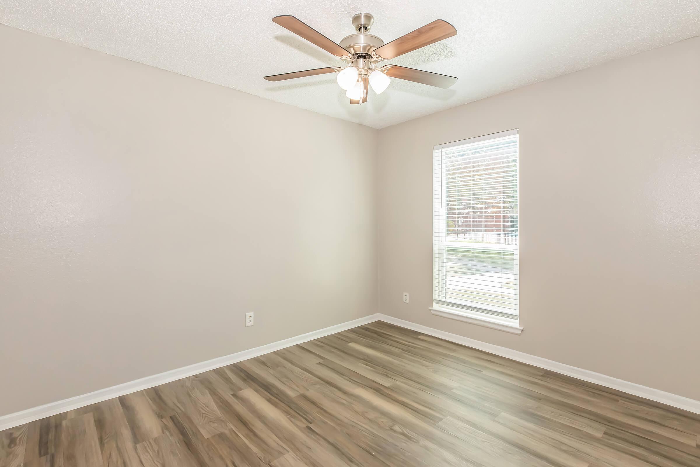 A light, airy room with beige walls and a ceiling fan. The floor is covered in wood-like laminate, and there is a window with white blinds allowing natural light to enter. The room appears spacious and unfurnished, suitable for various uses.