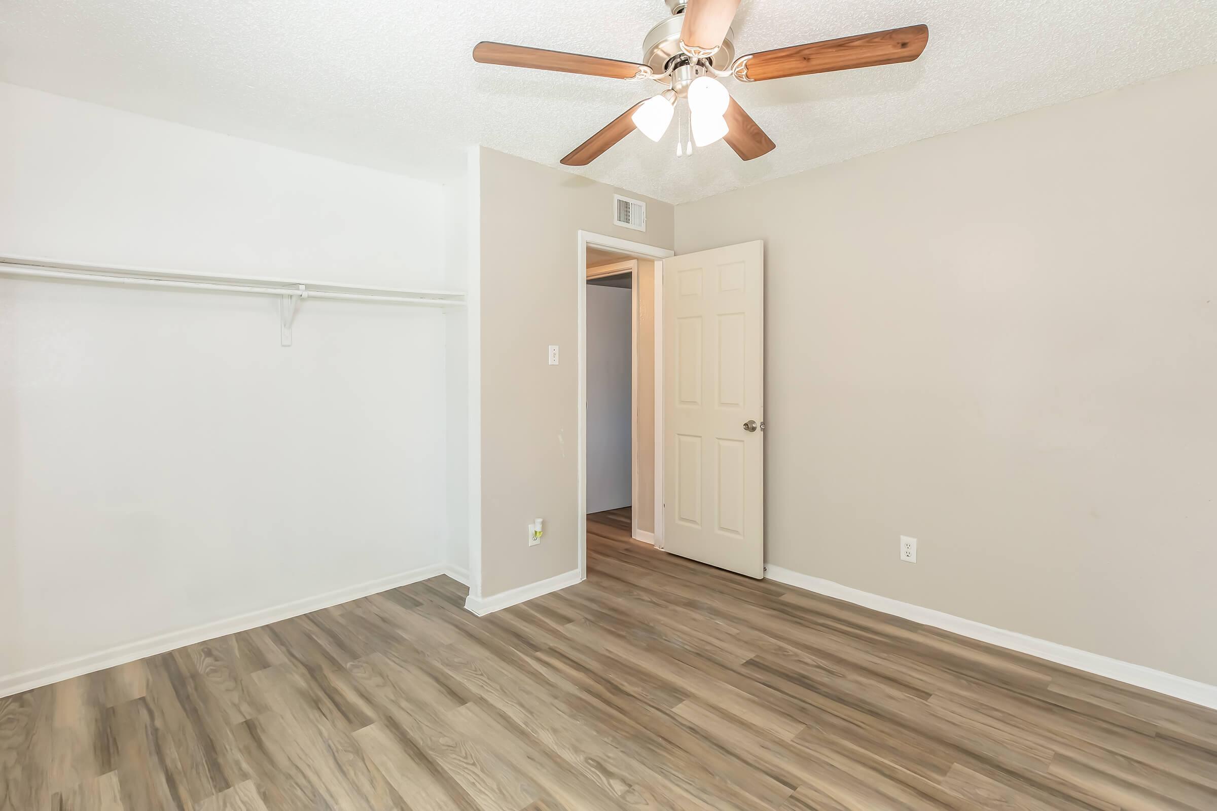 Interior of an empty room featuring light wood flooring, a ceiling fan with wooden blades, a closet with a single shelf, and a door leading to another space. The walls are painted a neutral color, creating a bright and open atmosphere.