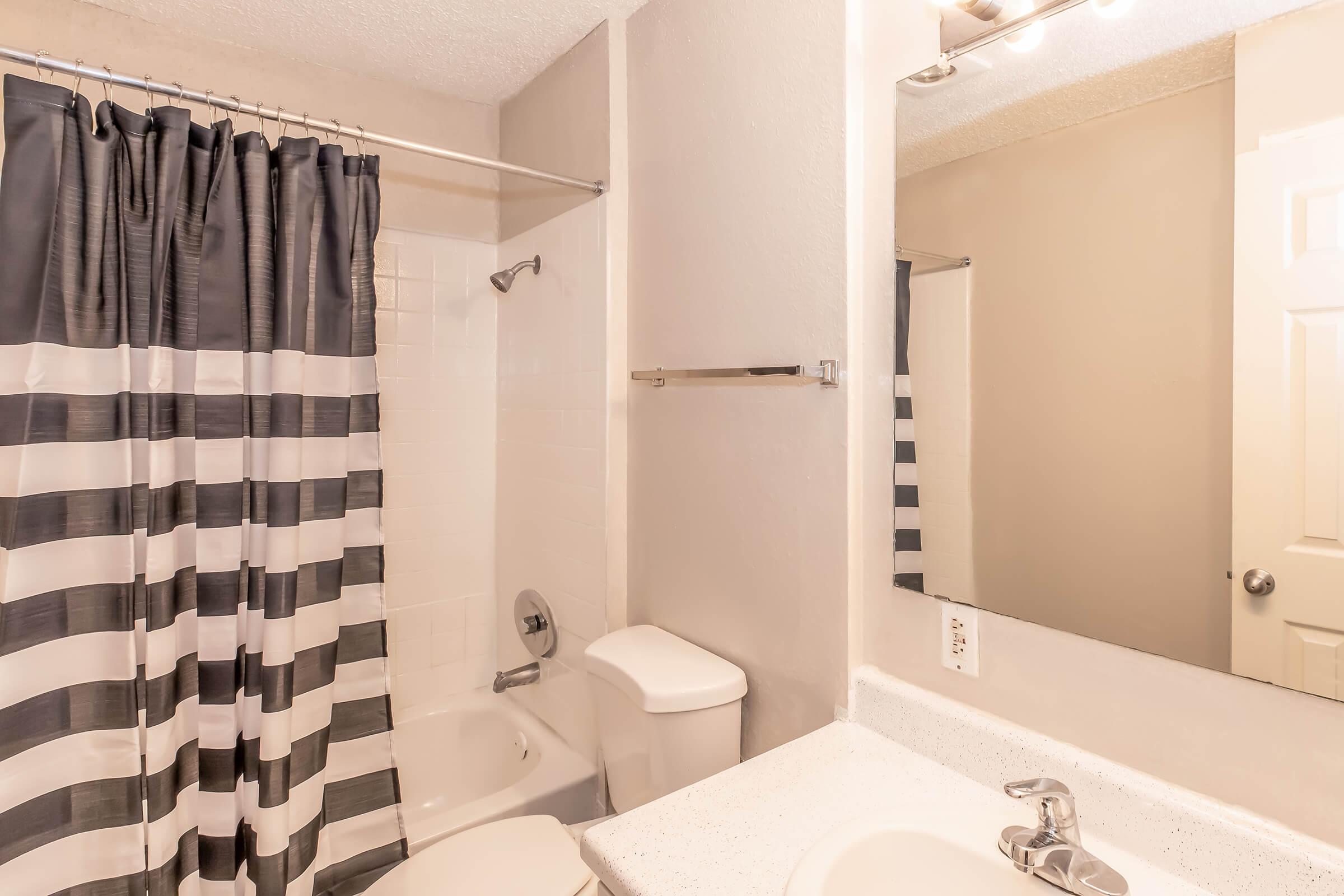 A bathroom featuring a striped black and white shower curtain, a bathtub with a shower head, a white toilet, and a vanity with a sink and faucet. A mirror is mounted above the sink, and light fixtures are positioned above the mirror. The walls are painted in a light color.