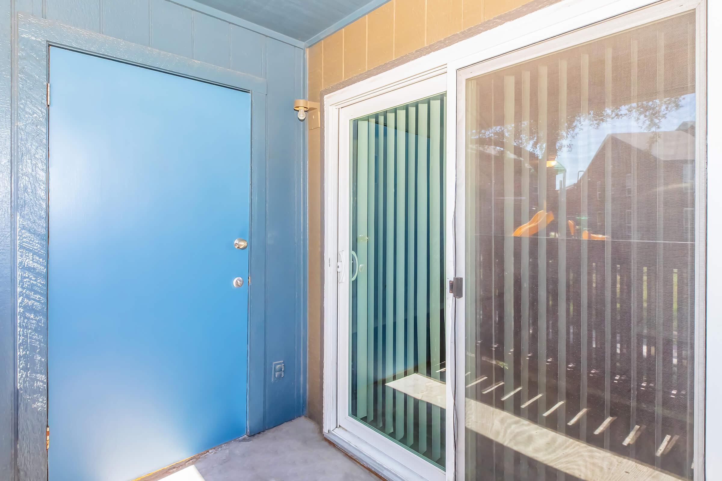 A blue door next to a sliding glass door with vertical blinds, leading to an outdoor area. The wall is a light brown color, and there is a concrete floor. Sunlight is illuminating the space, creating a bright atmosphere.