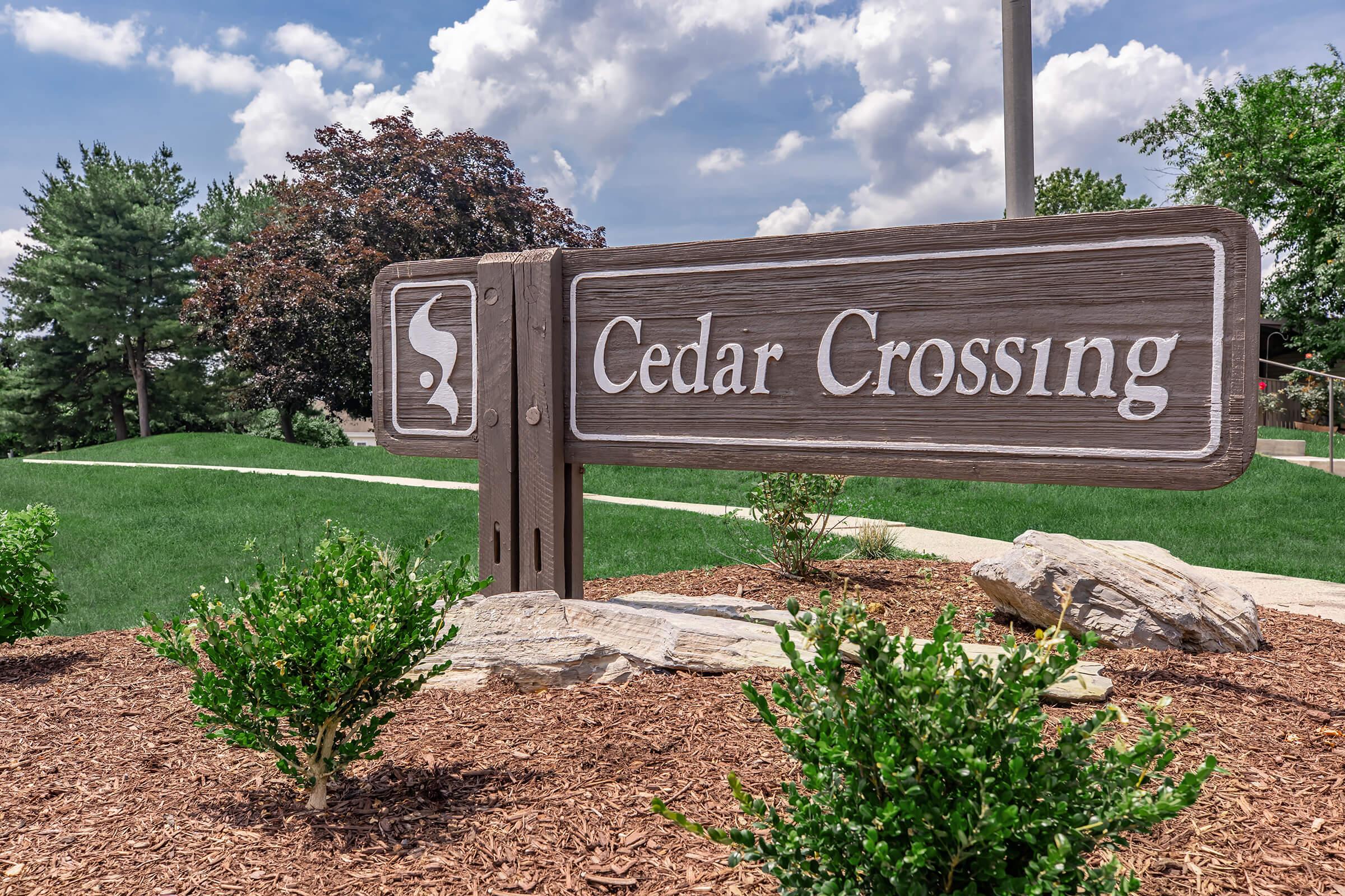 A wooden sign for "Cedar Crossing" set against a grassy background, featuring shrubs and trees. The sky is clear with some clouds, and a pathway is visible in the distance. The sign includes a stylized logo or symbol on the left side.