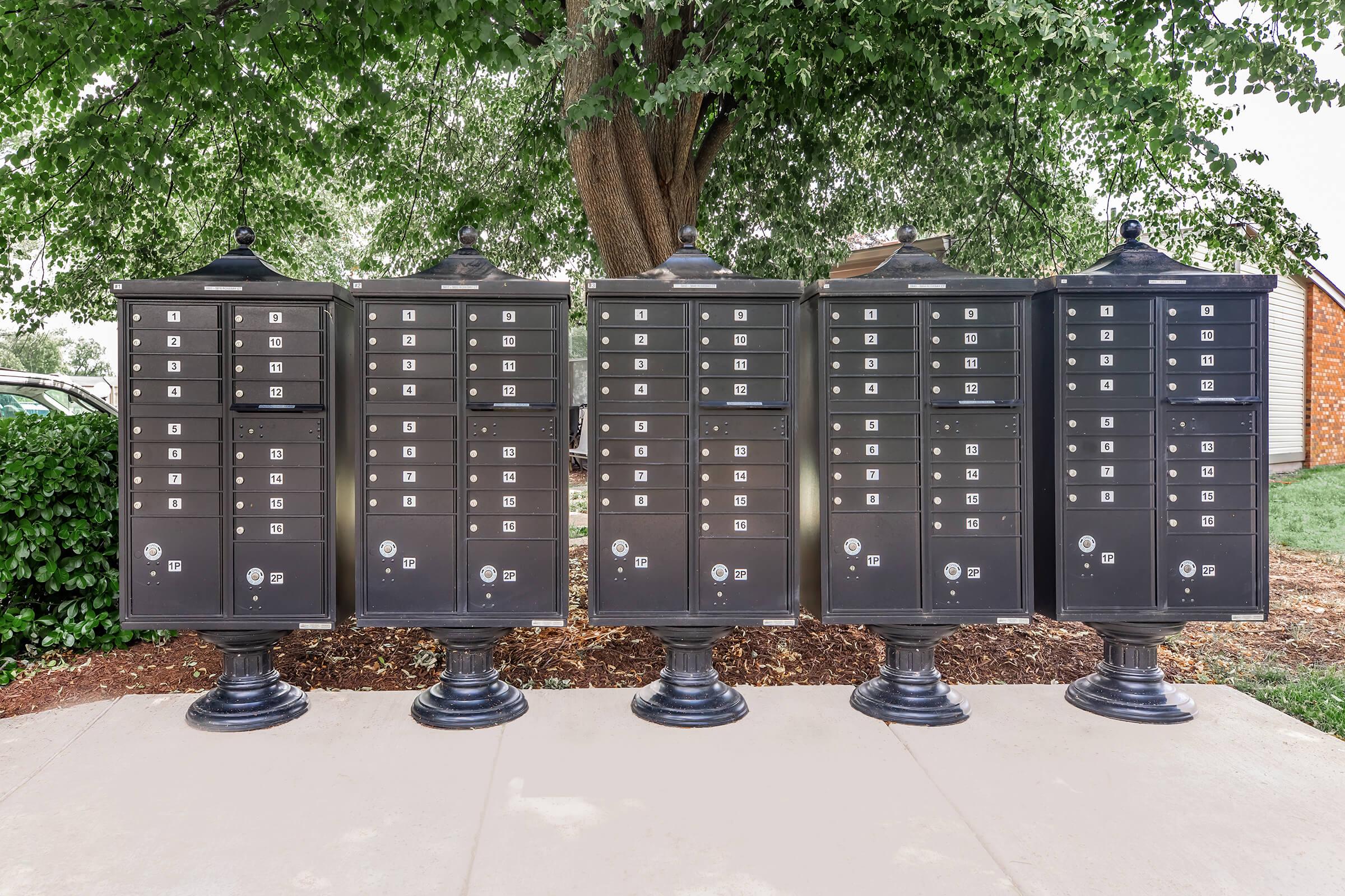 A row of five black residential mailbox units with multiple compartments for mail. Each mailbox features a decorative design and is mounted on a pedestal. The background includes greenery from a nearby tree and shrubs, with a paved walkway in front.