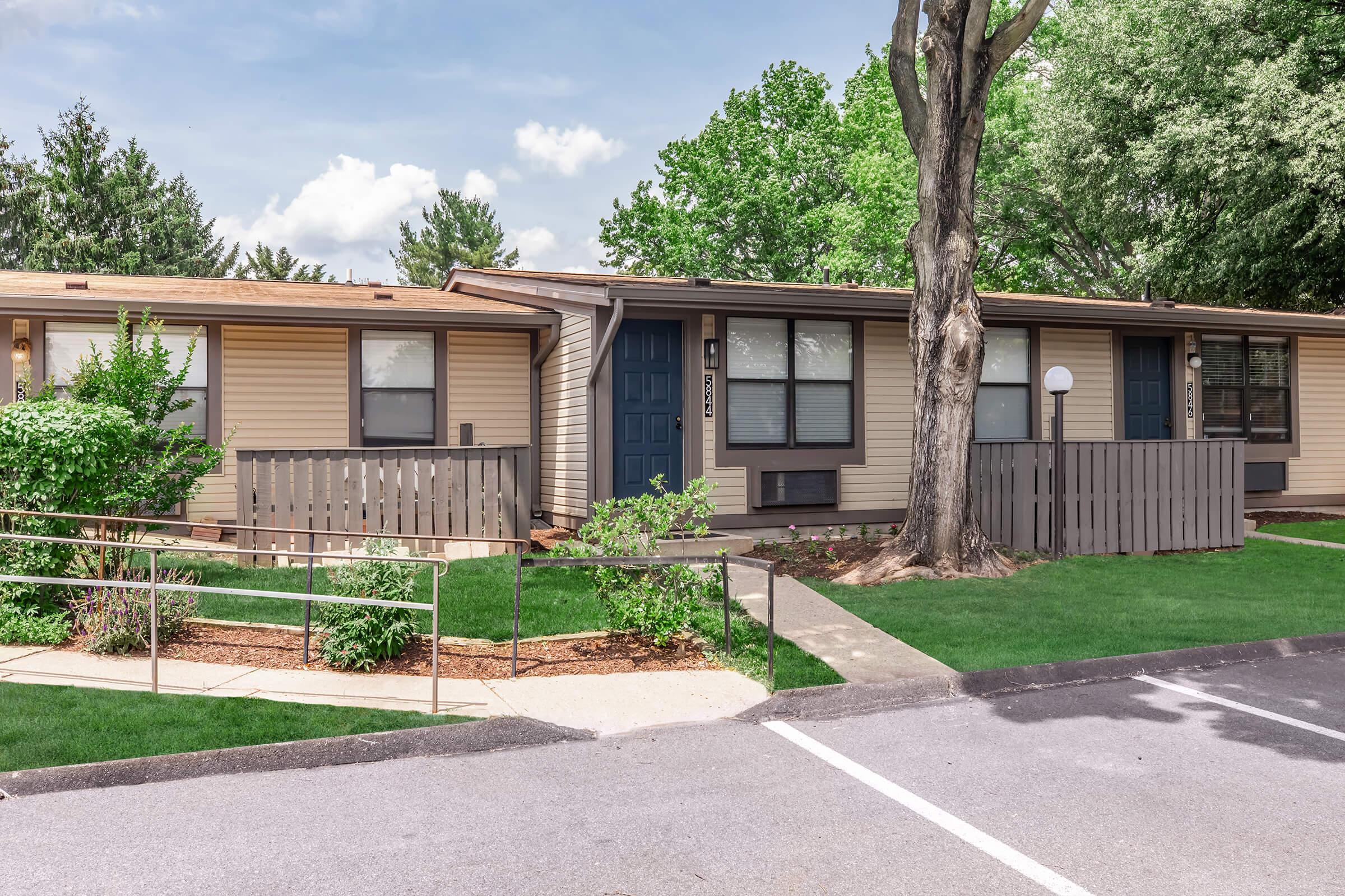 A row of residential buildings featuring a beige and brown exterior. Each unit has a front porch and a small garden area with shrubs. A paved pathway leads to the entrance, and a tree provides shade. The sky is partly cloudy, indicating a pleasant day.