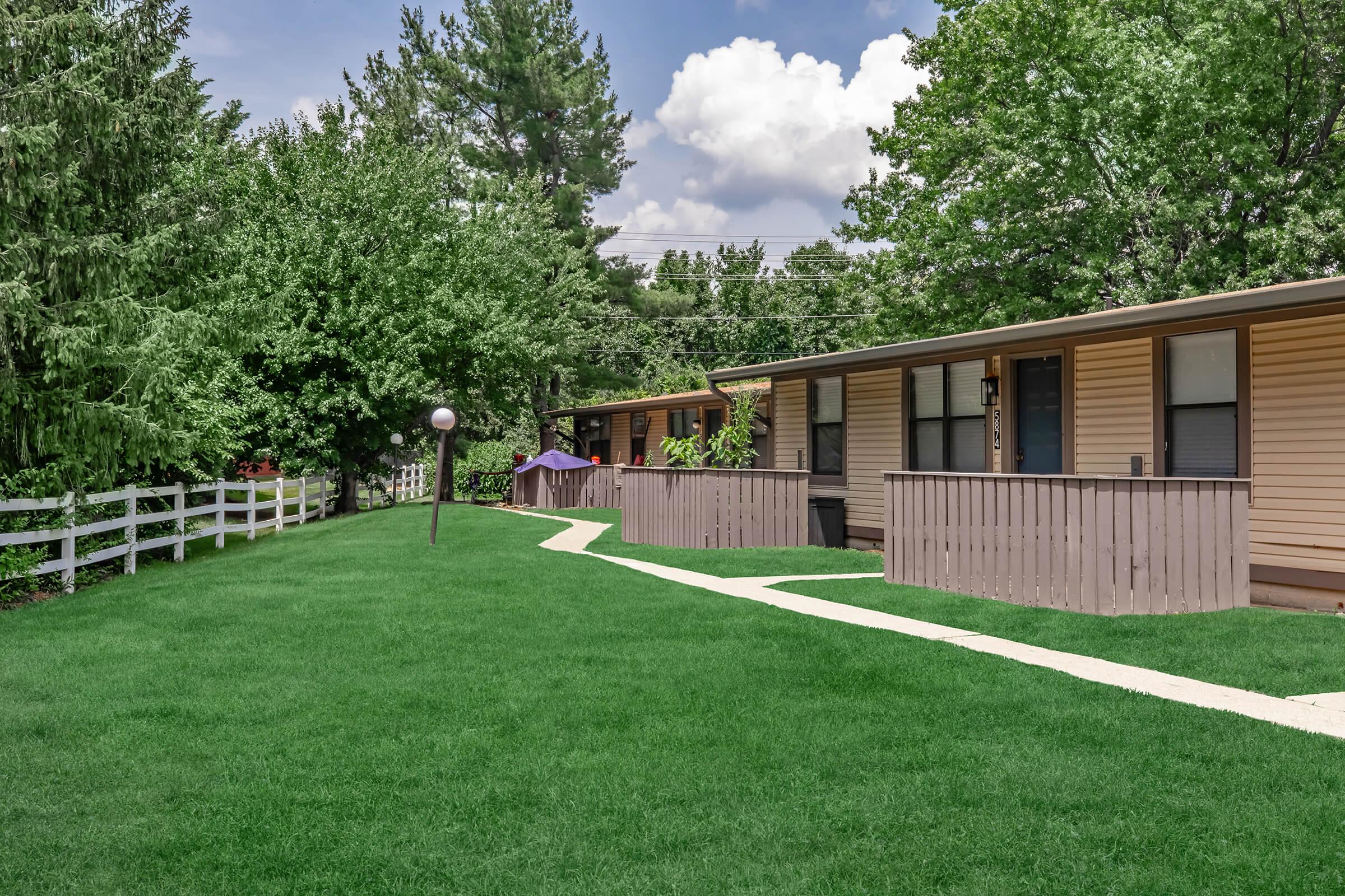 A well-maintained residential area featuring a neatly landscaped green lawn, bordered by a white picket fence. There are several single-story homes with light-colored siding, and trees surrounding the area under a partly cloudy sky. The pathway leads through the yard, enhancing the inviting atmosphere.