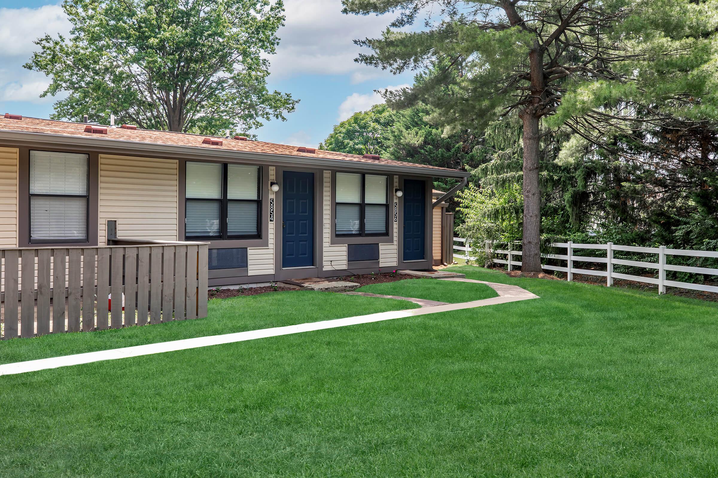 A view of a single-story residential building featuring two doors with blue accents, surrounded by lush green grass. A white fence runs along the right side, and trees are visible in the background, creating a serene, inviting atmosphere.