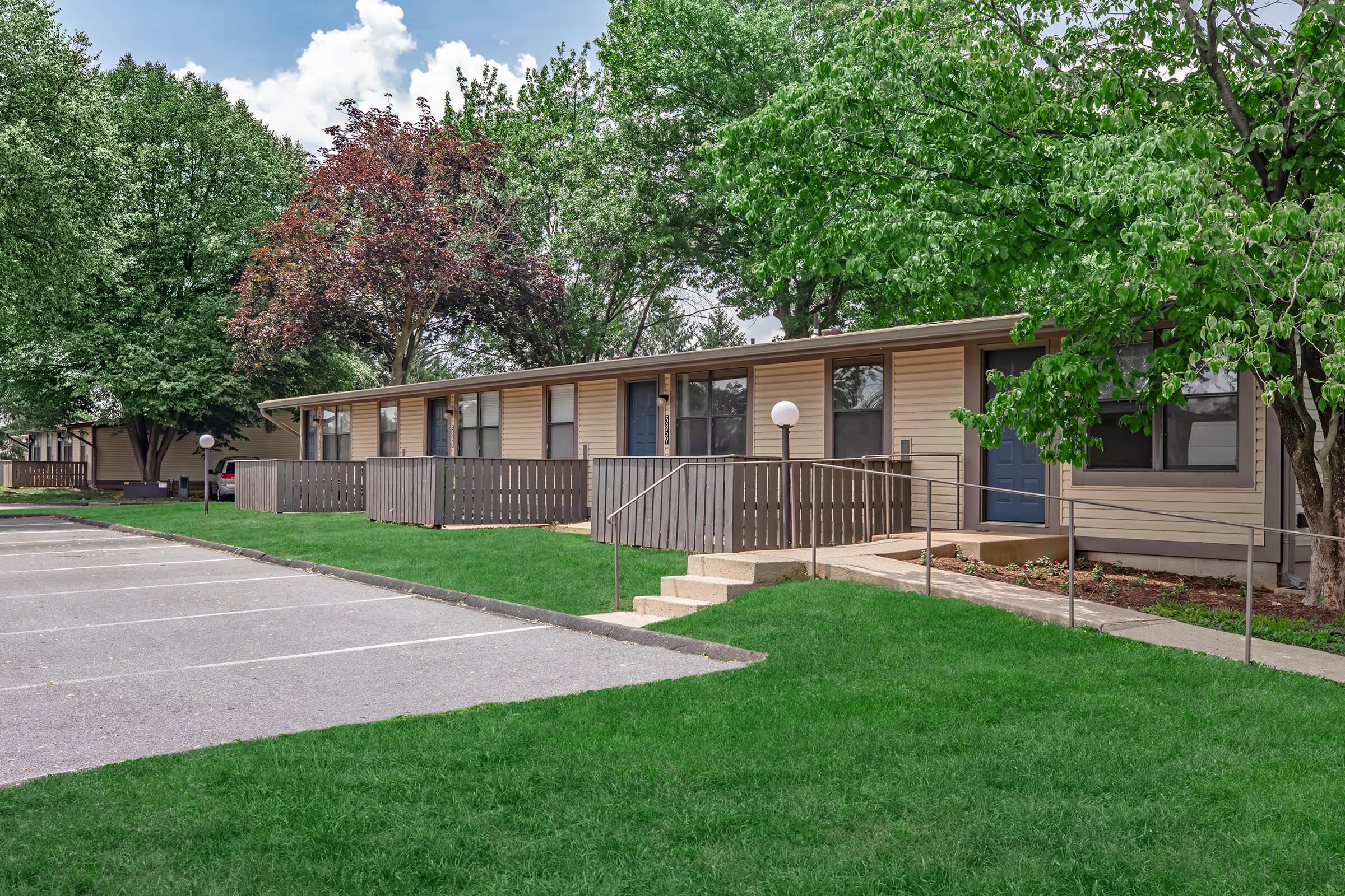 A row of single-story buildings surrounded by green grass and trees. Each unit has a small porch with railings, and there are light posts in front. A parking lot is visible in the foreground, with several empty parking spaces. The sky is partly cloudy, adding a bright atmosphere to the scene.