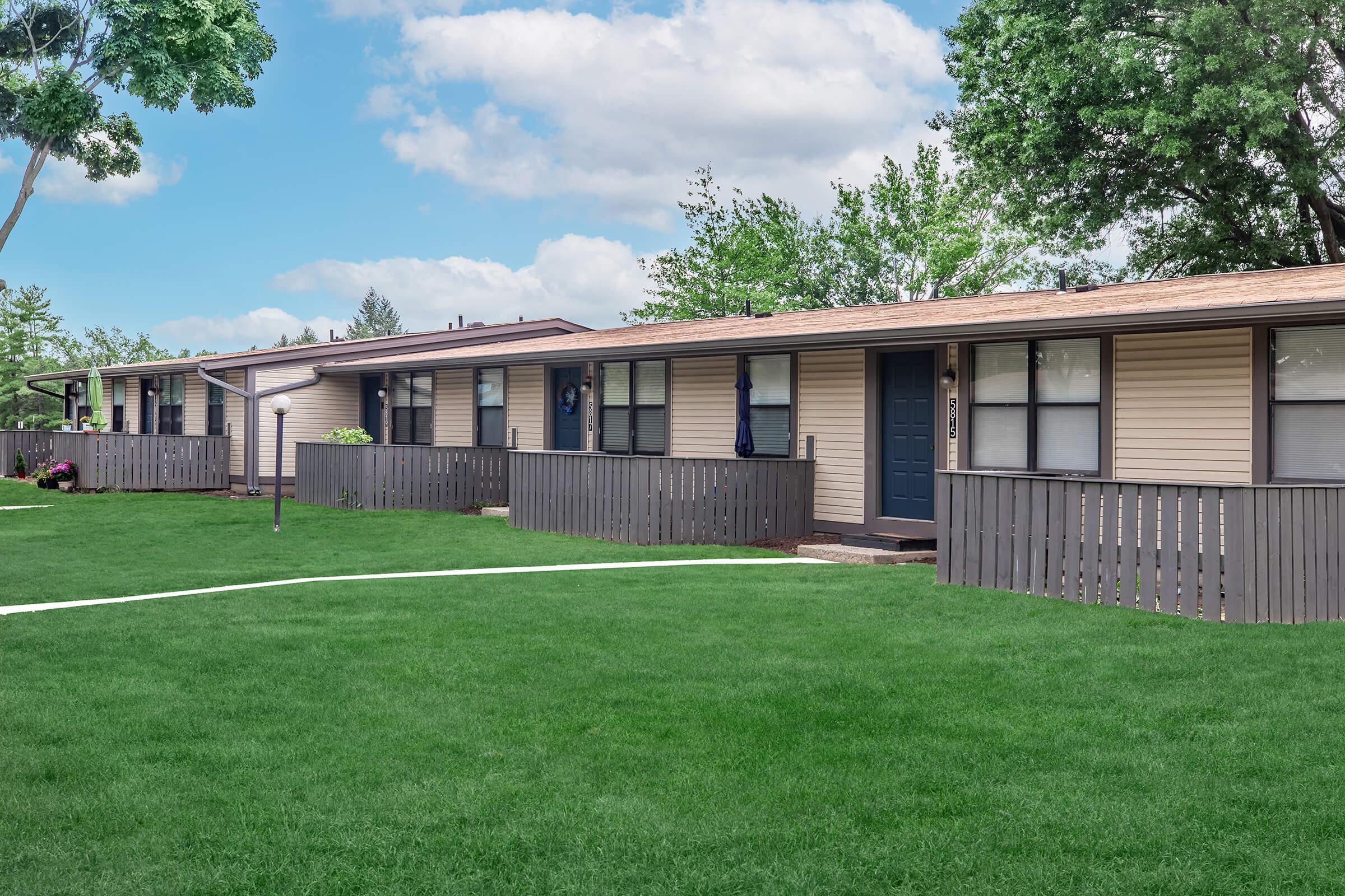 A row of single-story residential units with a green lawn in front. Each unit has a small porch and a wooden fence separating them. There are trees in the background and a partly cloudy blue sky above, creating a serene suburban atmosphere.