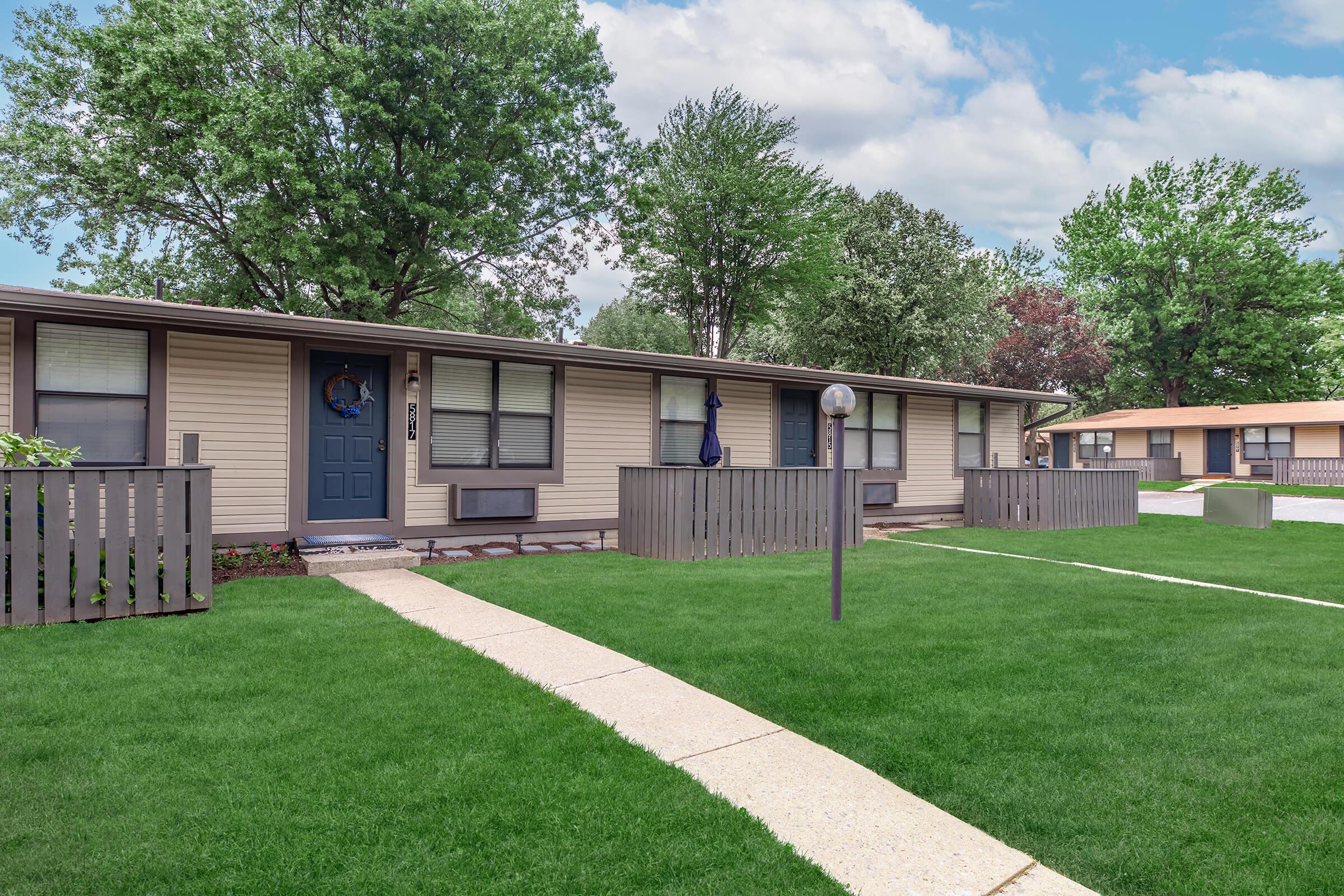 A well-maintained row of single-story apartments, featuring green lawns and simple wooden fences. Each unit has a door, with one adorned by a decorative wreath. Trees can be seen in the background under a partly cloudy sky. A concrete pathway leads through the grassy area, creating a neat, inviting atmosphere.