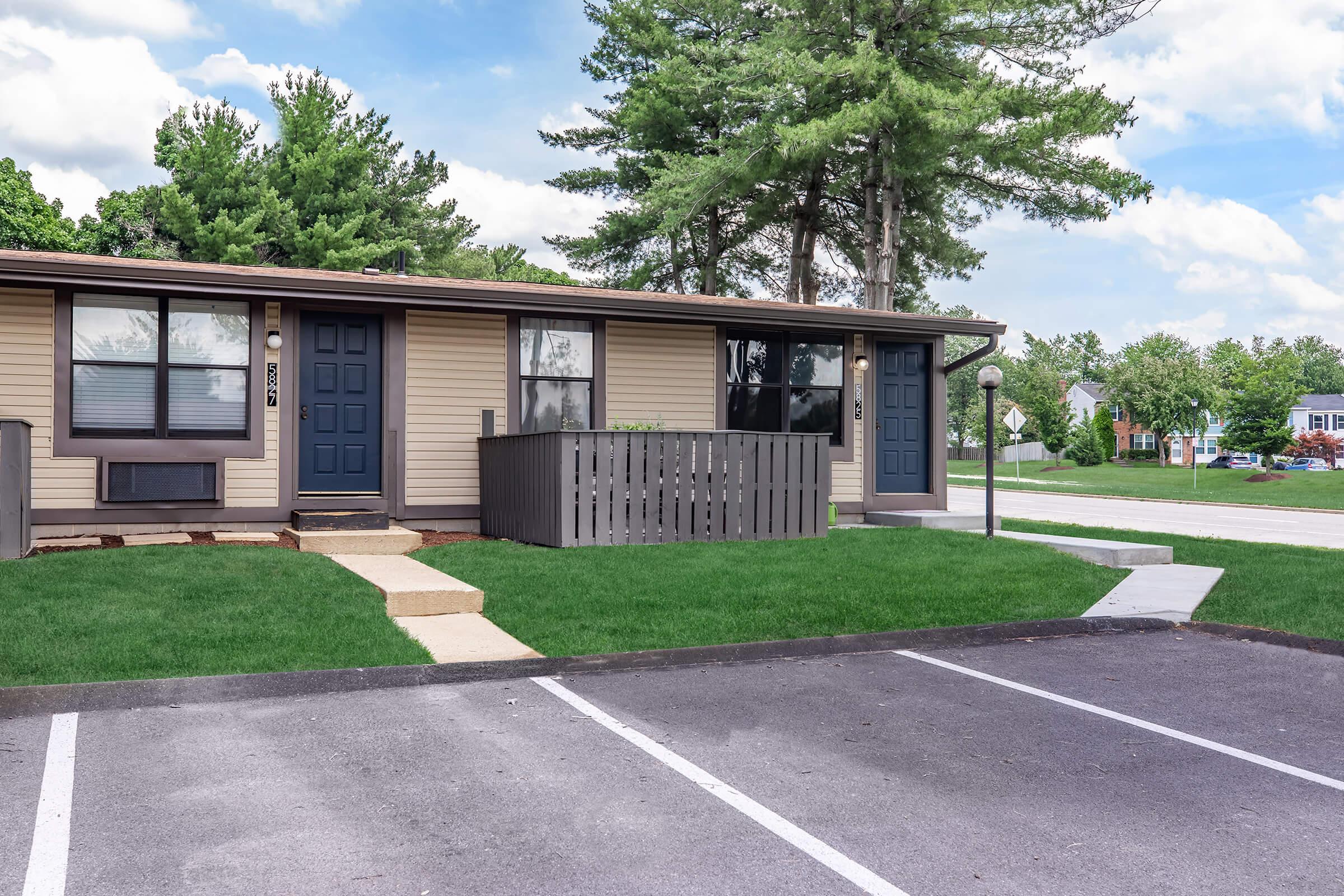 A two-story apartment building with light brown siding and dark blue doors, surrounded by green grass and small trees. A gray wooden fence is visible in front of the doors. The parking lot has clearly marked spaces, and there is a sidewalk leading to the entrance. The sky is partly cloudy.